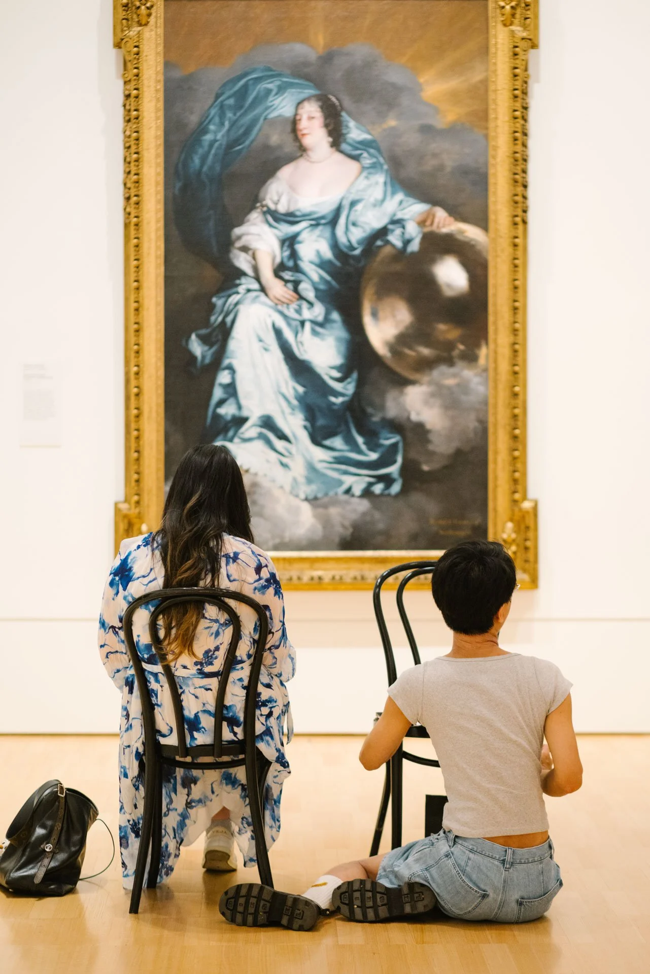 Three people observe a large portrait of a woman in a historical painting at an art museum. One person is sitting on the floor, another is seated in a chair, and a third has long hair and is wearing a floral dress.