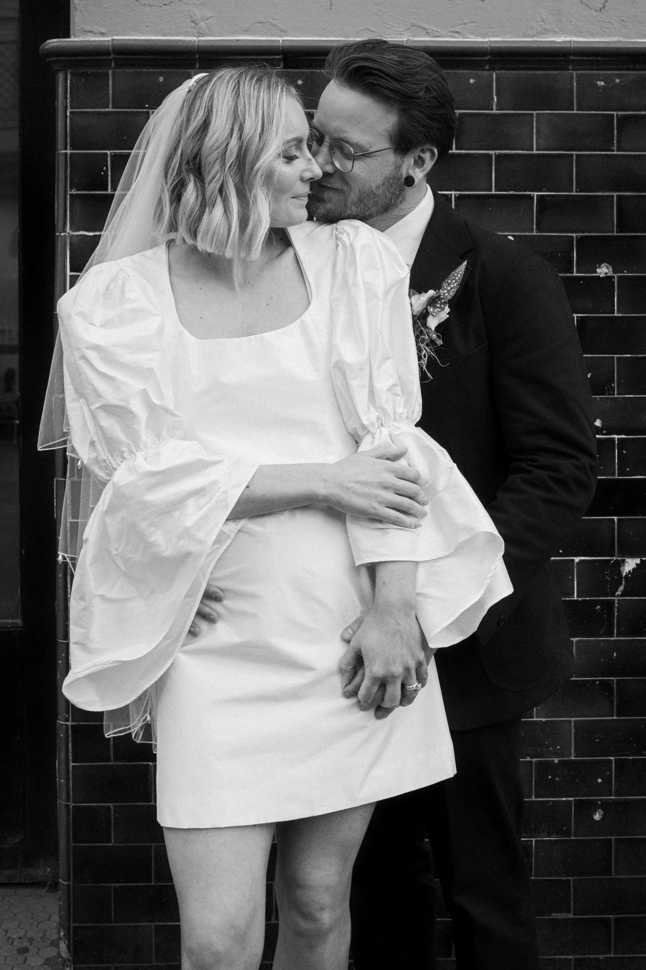 A couple, dressed in wedding attire, sharing an intimate moment while sitting against a brick wall. The woman has shoulder-length blonde hair and a veil; the man has dark hair, glasses, and earrings. They are close, with noses touching, and the man h