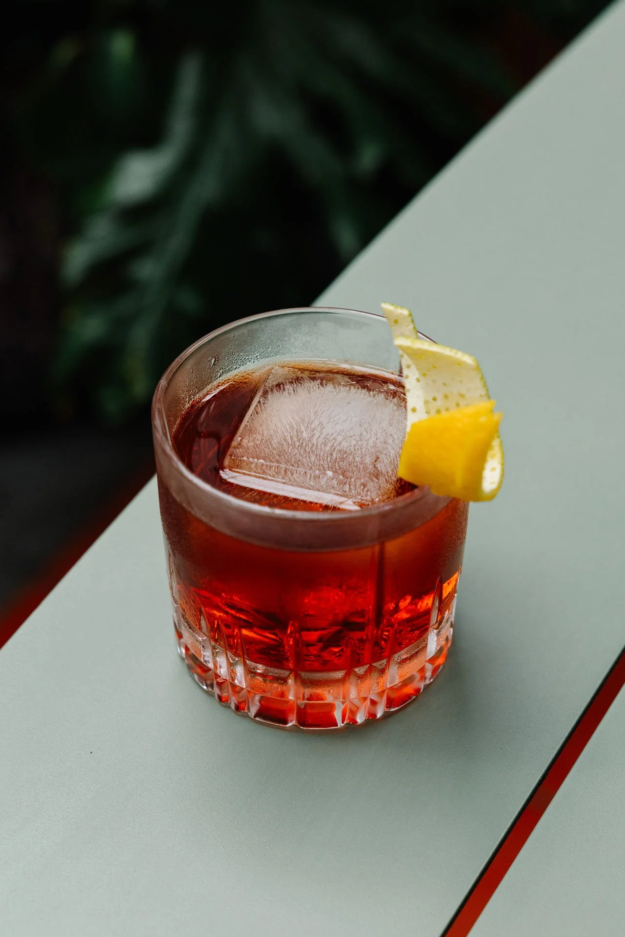 A glass of red beverage with ice cubes and a lemon wedge garnish placed on a light-colored surface.
