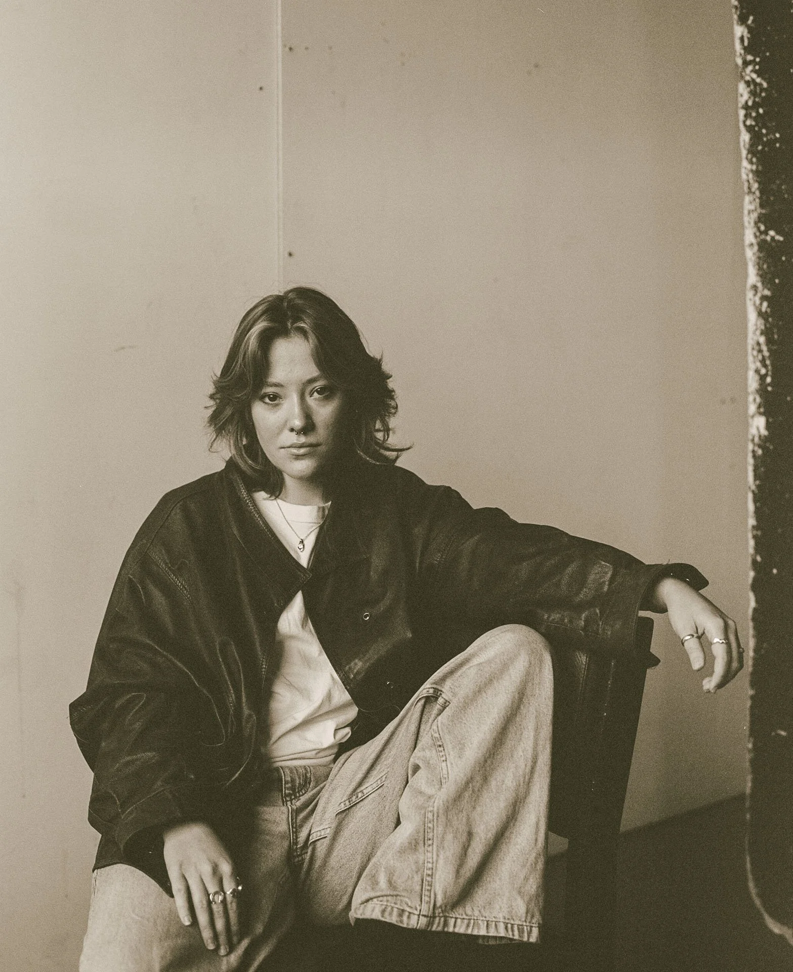 A woman with shoulder-length hair and multiple rings sitting on a black leather chair, wearing a black jacket over a white shirt and light-colored jeans, looking at the camera.