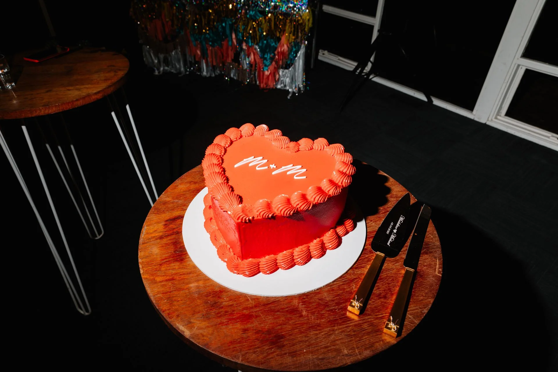 A heart-shaped cake with red frosting and decorative piping, placed on a white cake board on a round wooden table at a celebration event.