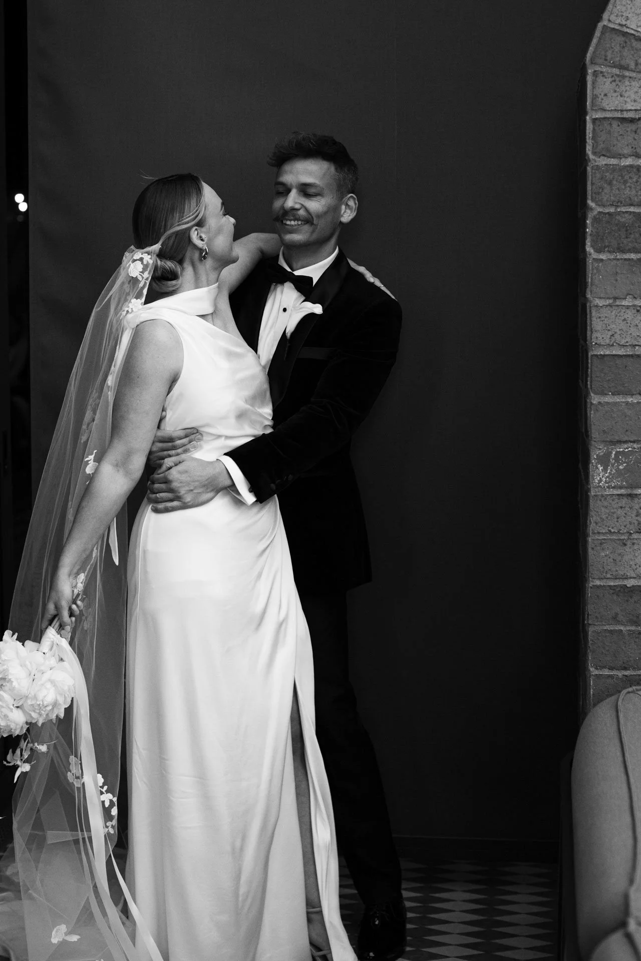 Black and white photo of a happy bride and groom sharing a moment, the bride in a wedding dress with a veil and the groom in a tuxedo, standing against a dark background and brick wall.