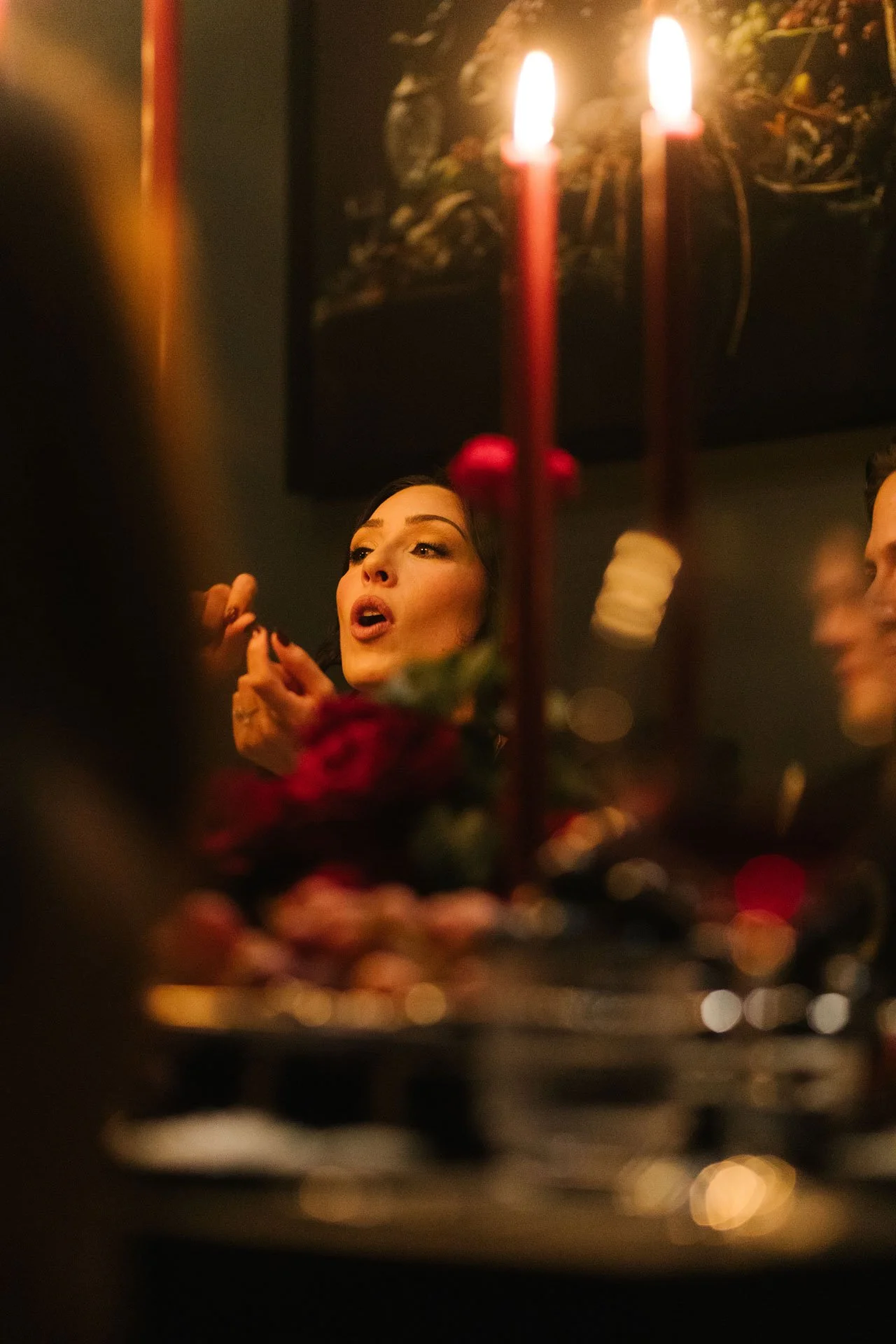 A woman with dark hair and makeup appears to be speaking or singing in a dimly lit setting. Two lit candles are in the foreground, and blurred decorative items are visible in the background.