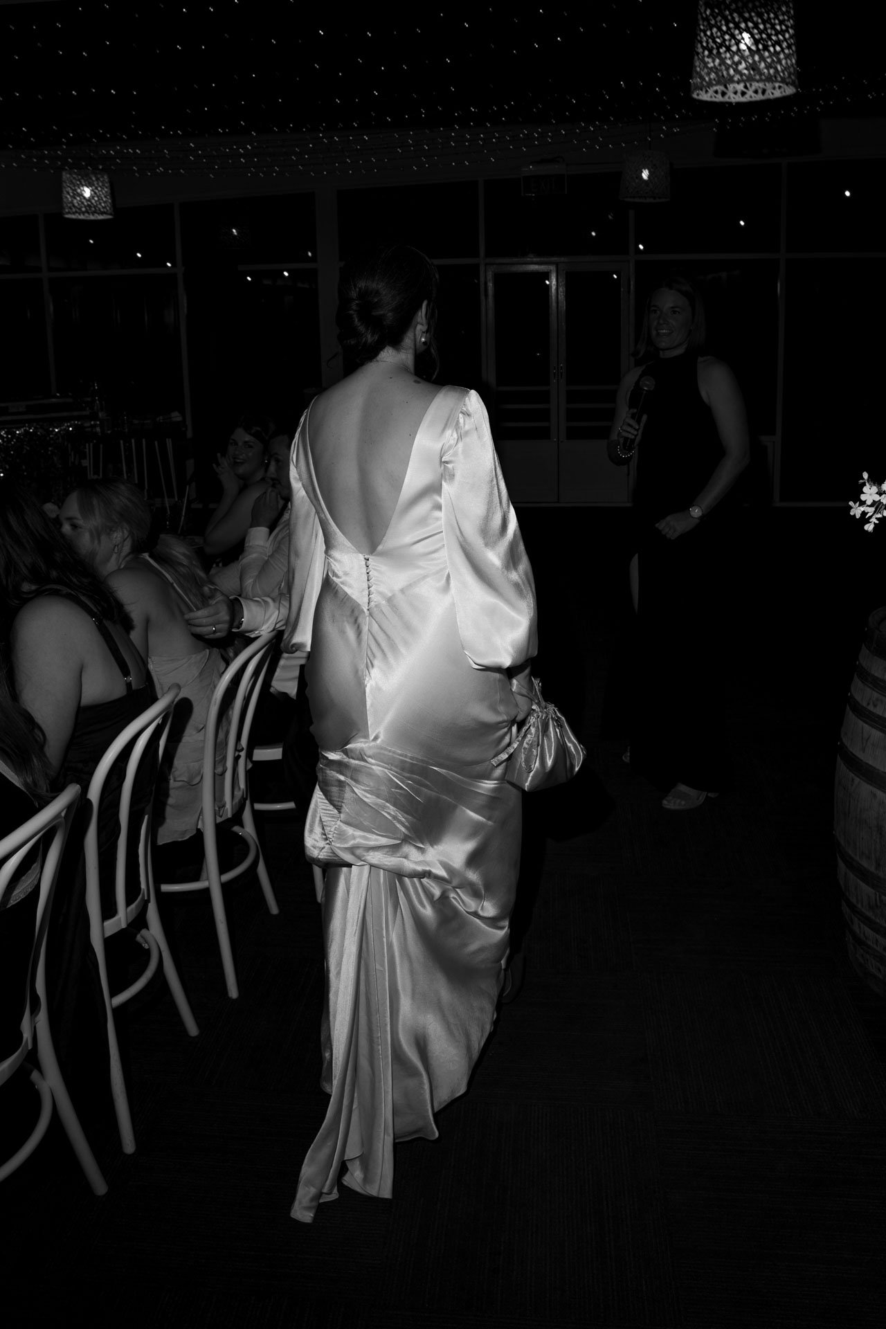 A woman in a satin wedding gown with a deep back, standing in a room with several people seated at tables, a woman with a microphone speaking, and a ceiling decorated with string lights.