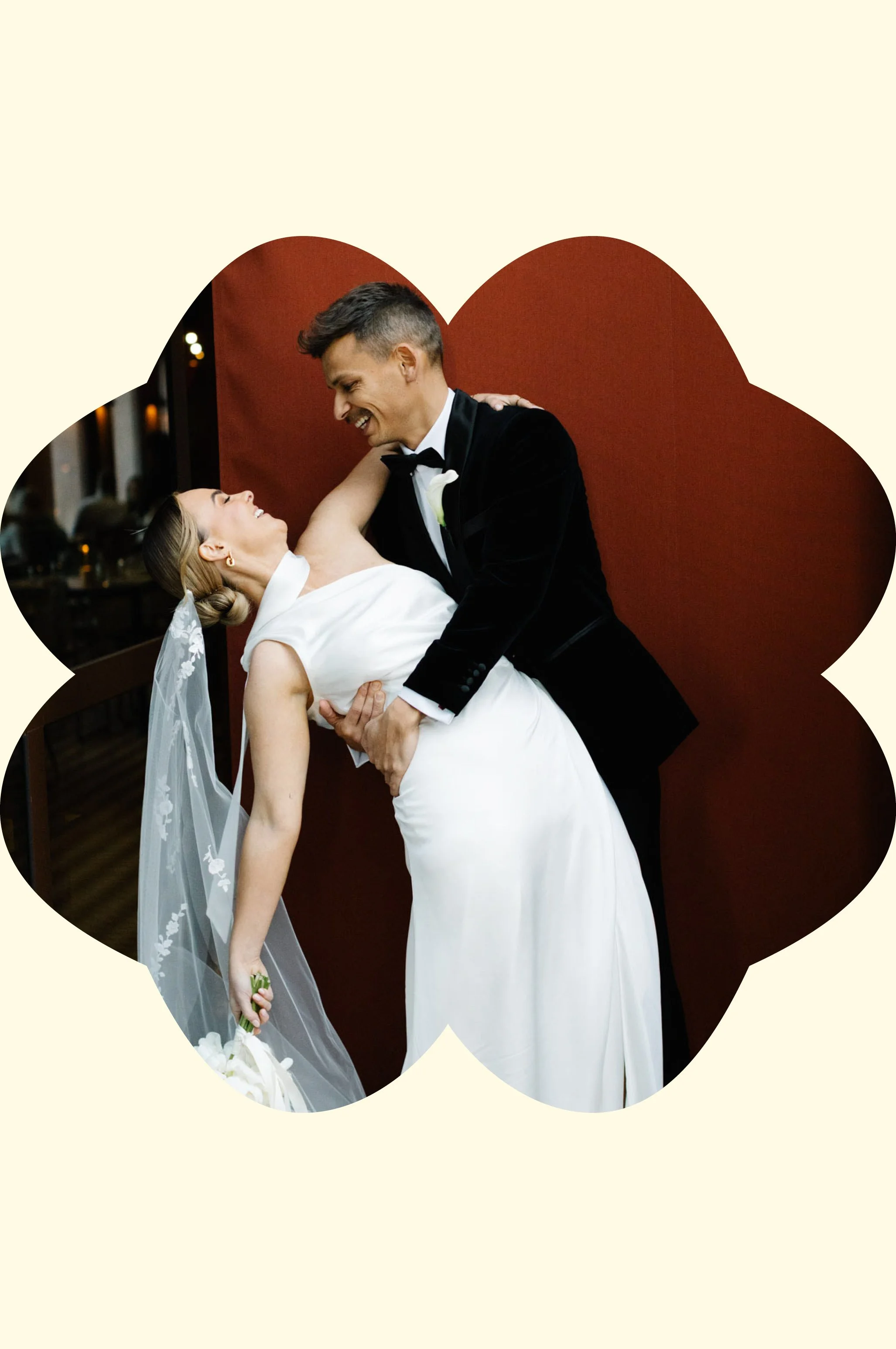 A bride and groom dance during their wedding reception. The groom is dressed in a black tuxedo and the bride is wearing a white wedding gown and veil. They are smiling and looking at each other affectionately.
