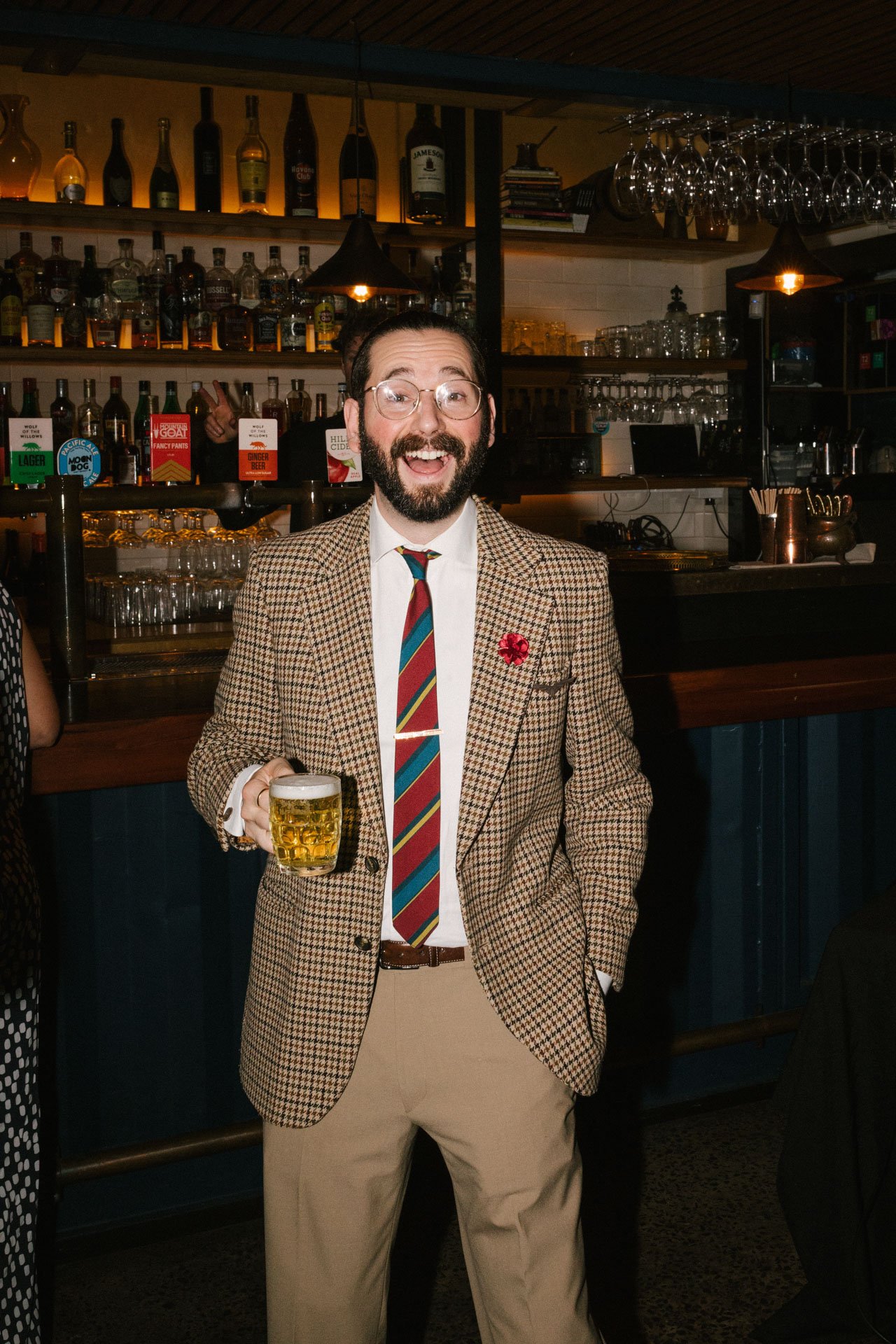 A man with glasses and a beard smiling while holding a glass of beer in a bar with shelves of bottles behind him.
