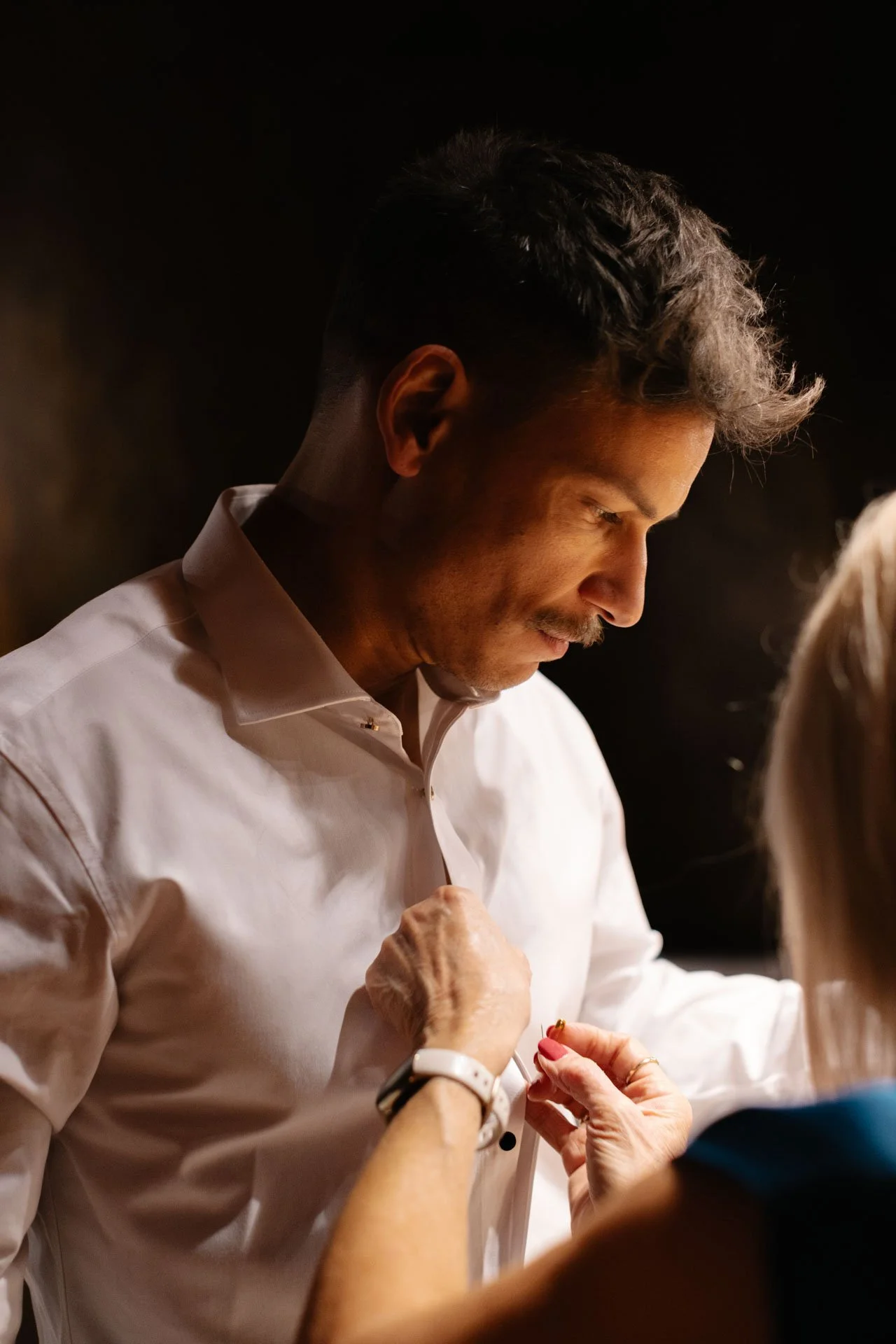 A man in a white shirt having a ring put on his finger by a woman. The man has short dark hair and is looking downward, in a dimly lit setting.