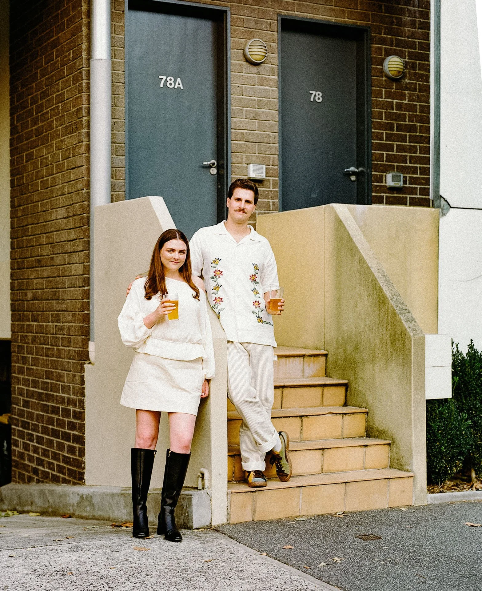 A man and woman standing on stairs outside a brick building, holding drinks, with two doors labeled 78A and 78 in the background.