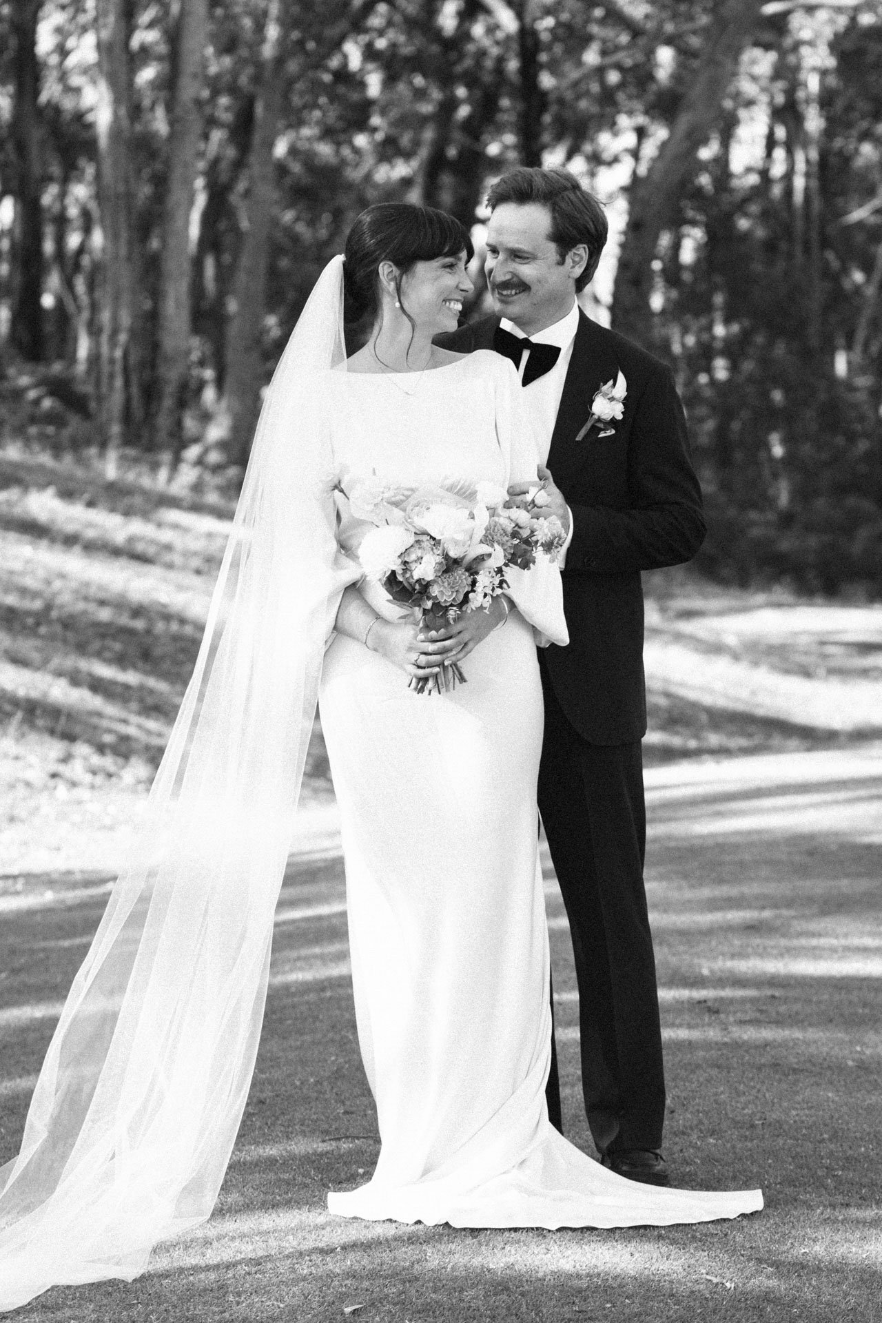 A black and white photo of a bride and groom smiling at each other in an outdoor setting with trees in the background. The bride is holding a bouquet and wearing a wedding dress with a veil, while the groom is dressed in a tuxedo with a boutonniere.