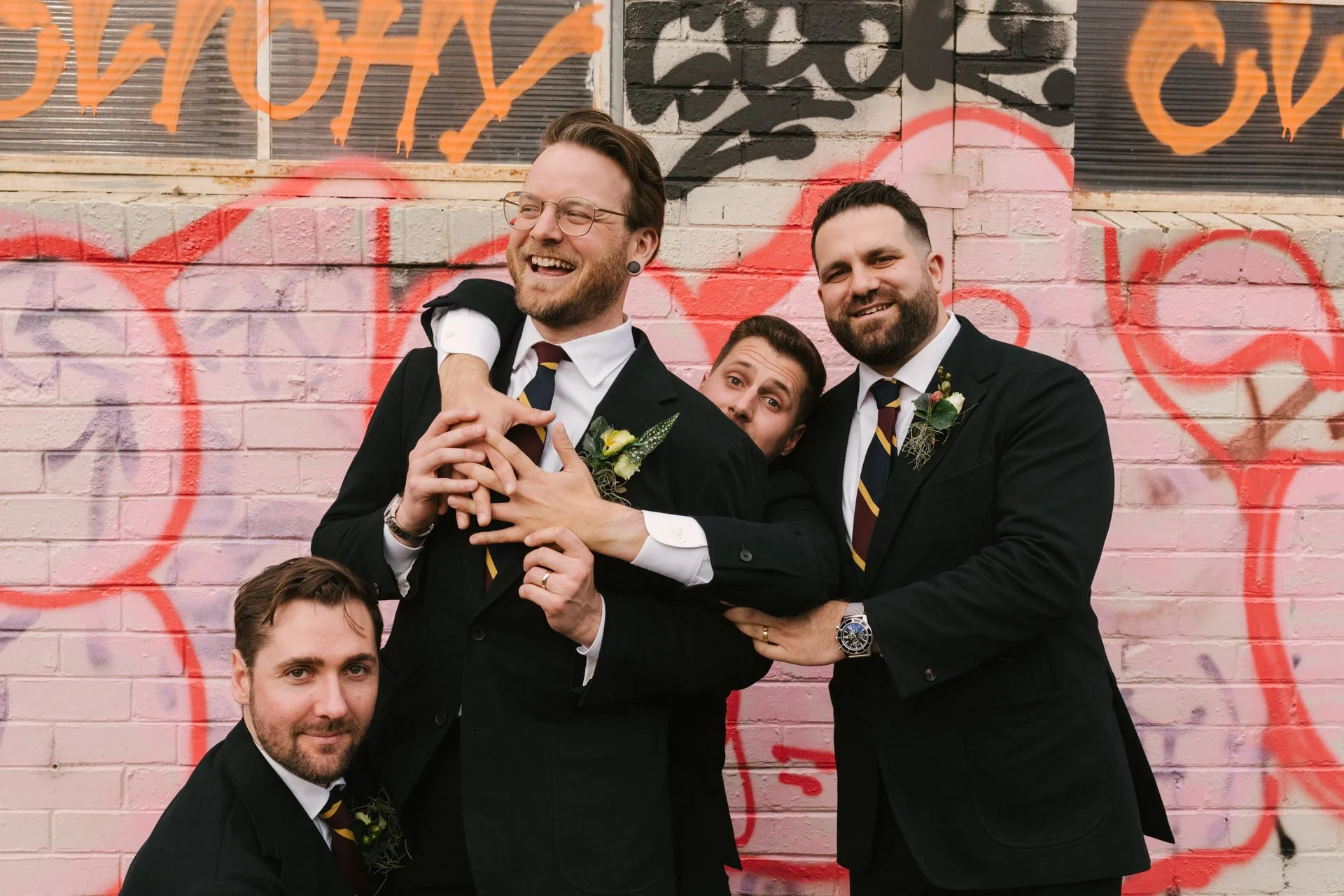 Four men dressed in suits and ties smiling and posing in front of a pink brick wall with graffiti art.