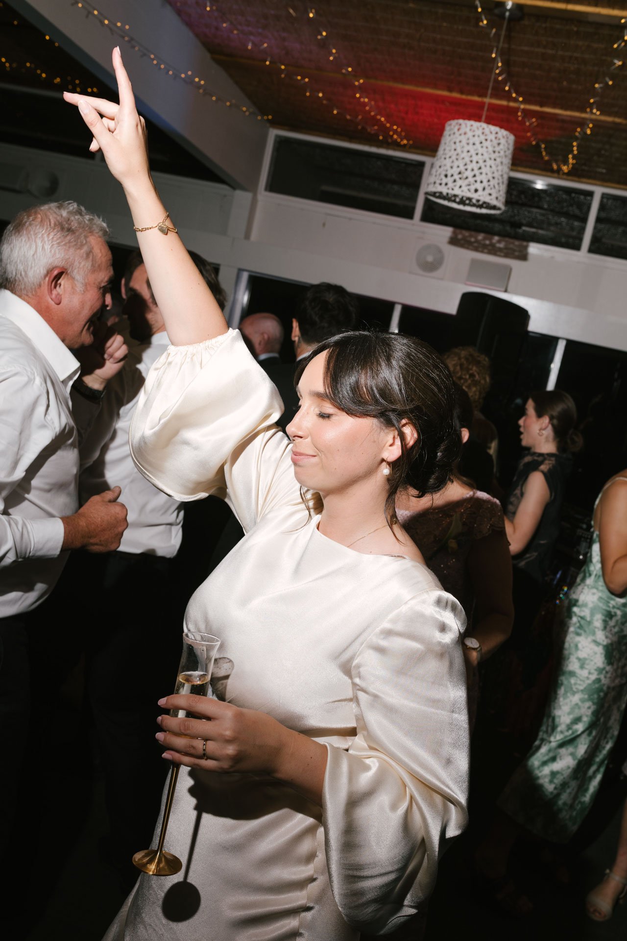 A woman with dark hair in an elegant white satin dress holding a champagne glass at a party or celebration, with multiple people dancing and socializing in the background, under string lights and a decorative ceiling light.