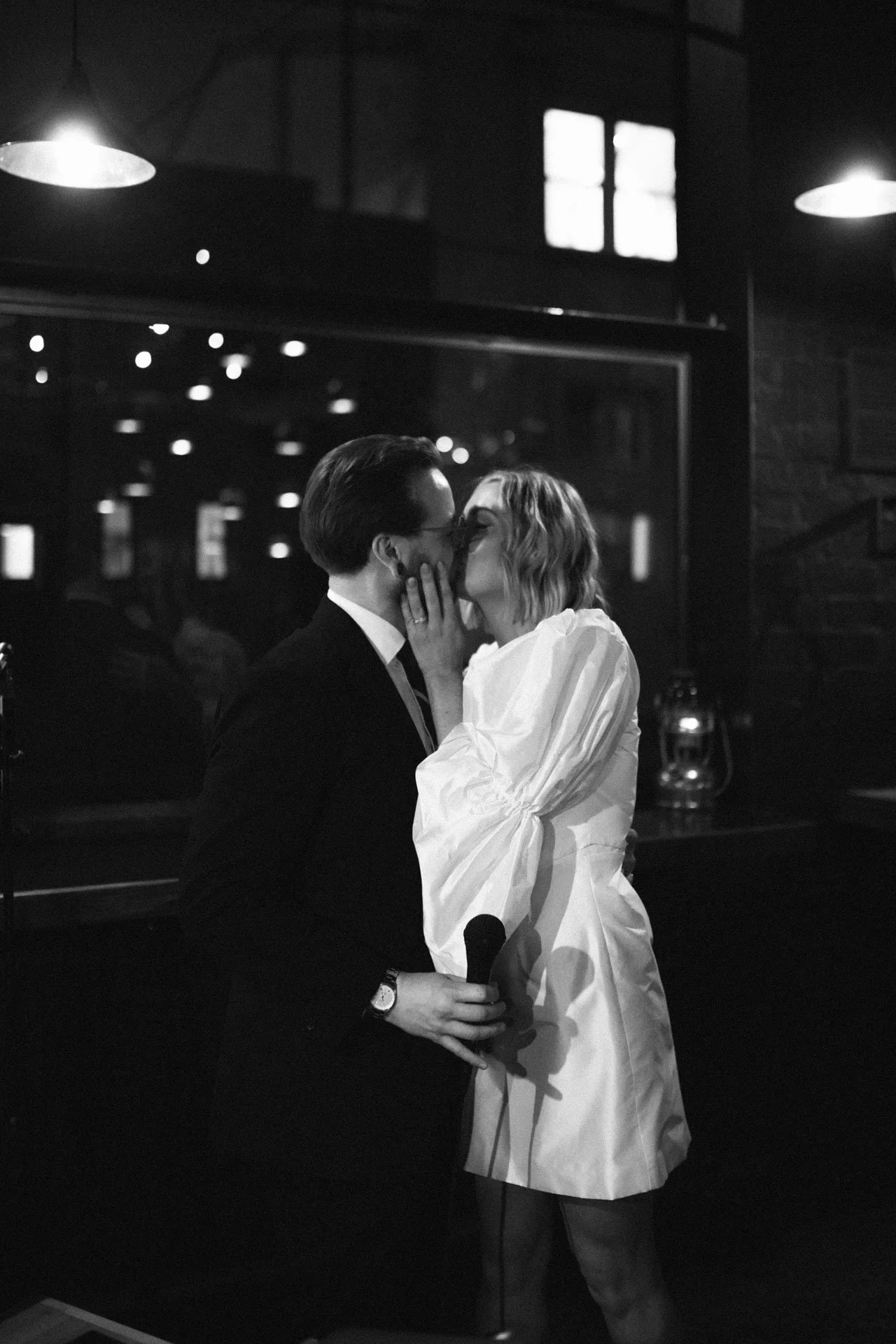 A man and woman share a kiss in a dimly lit indoor space, with the man holding a microphone and wearing a suit, while the woman is dressed in a white blouse.