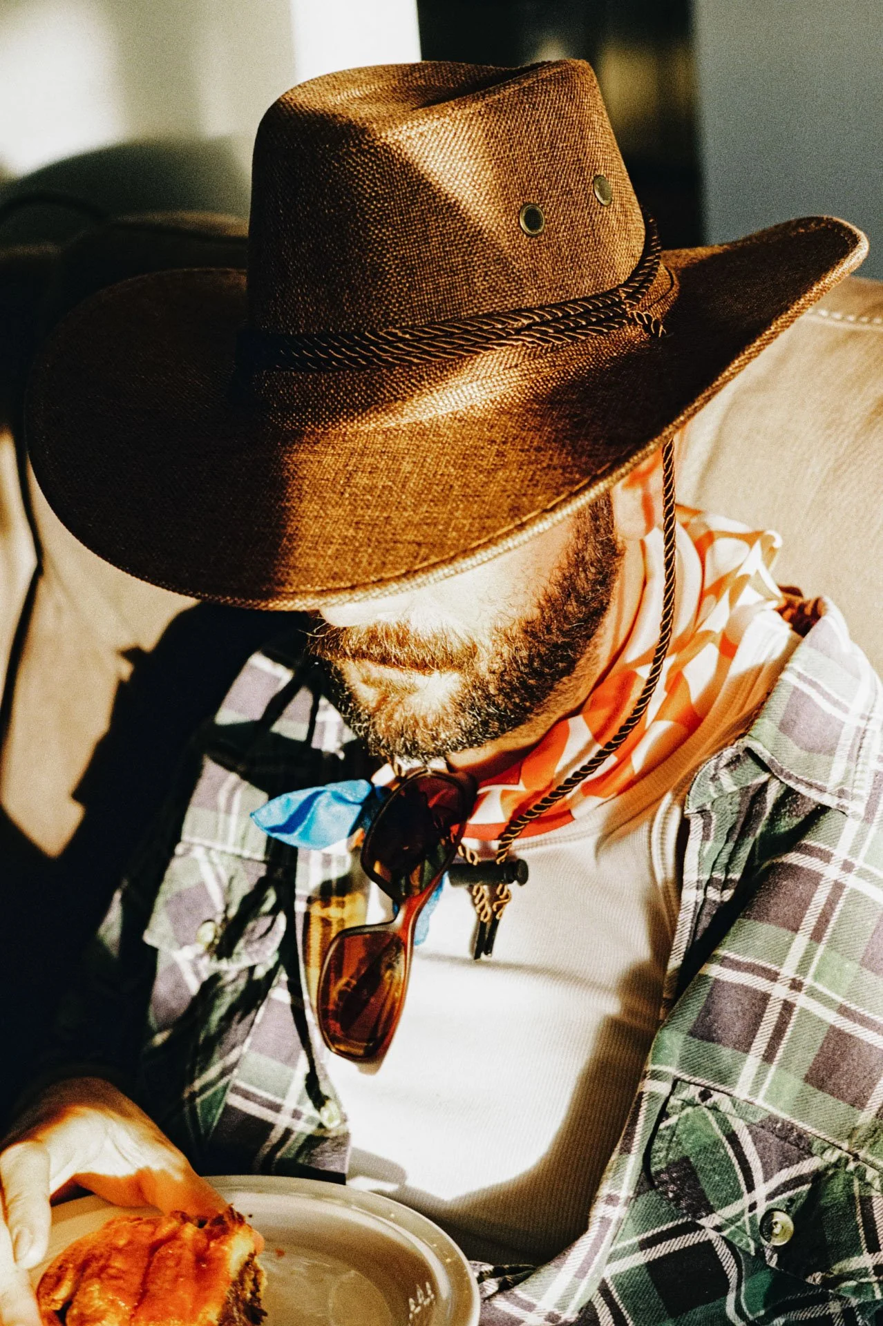A man wearing a large cowboy hat with eyelets, a plaid shirt, sunglasses hanging from his neck, and a scarf. He is sitting at a table with a plate of pizza.