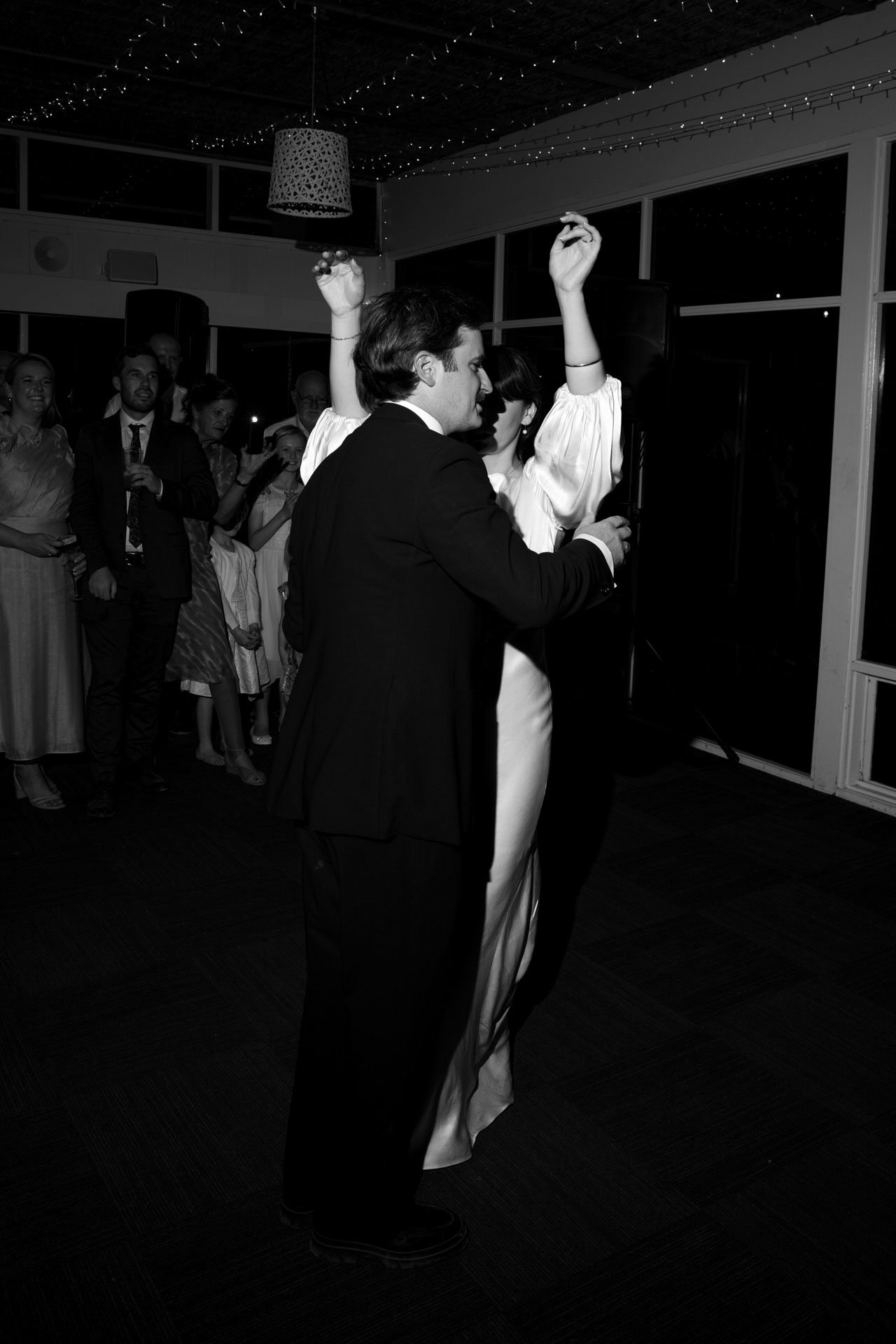 Black and white photo of a wedding reception with a bride and groom dancing, surrounded by guests. The bride is wearing a white dress and the groom is in a suit.