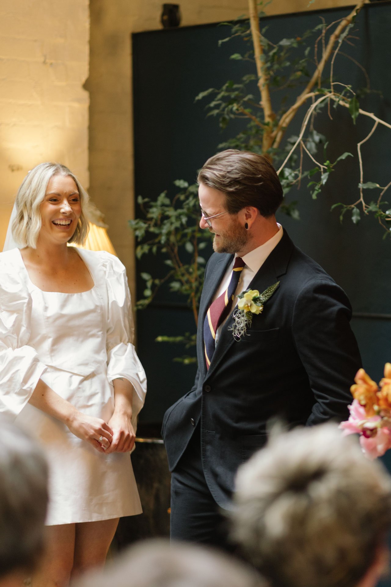 A bride and groom during their wedding ceremony, smiling and facing each other indoors.