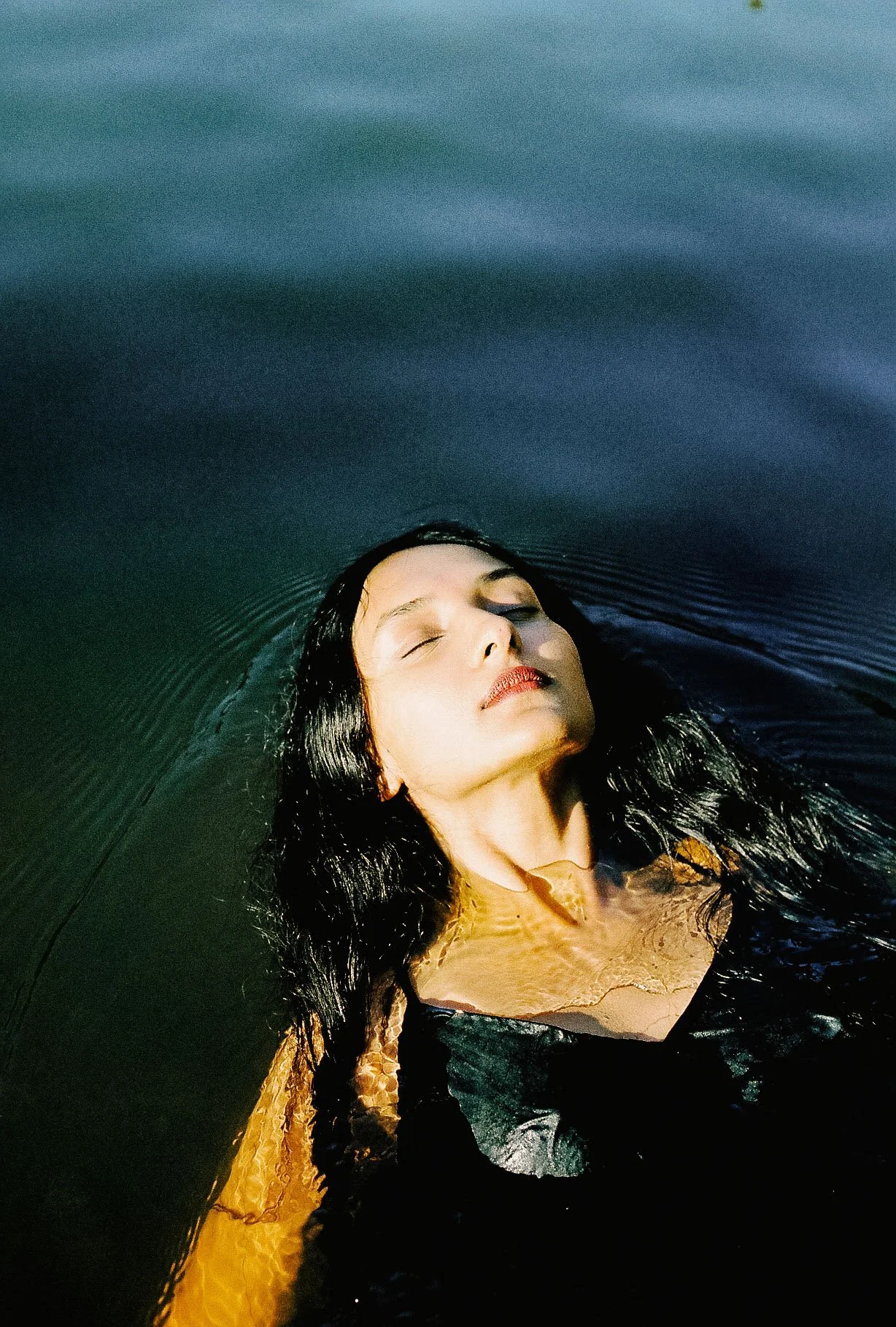 A woman with closed eyes and wet hair floating in dark water with her head tilted back and face up, showing a serene expression.