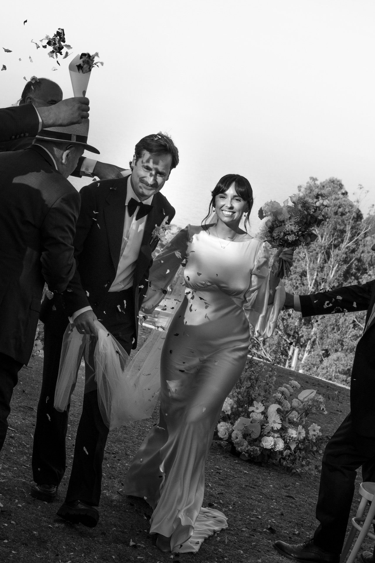 Black and white photo of a bride in a satin wedding dress walking outdoors, smiling, with a bouquet of flowers, being celebrated by friends or family throwing confetti, some holding cones with confetti.