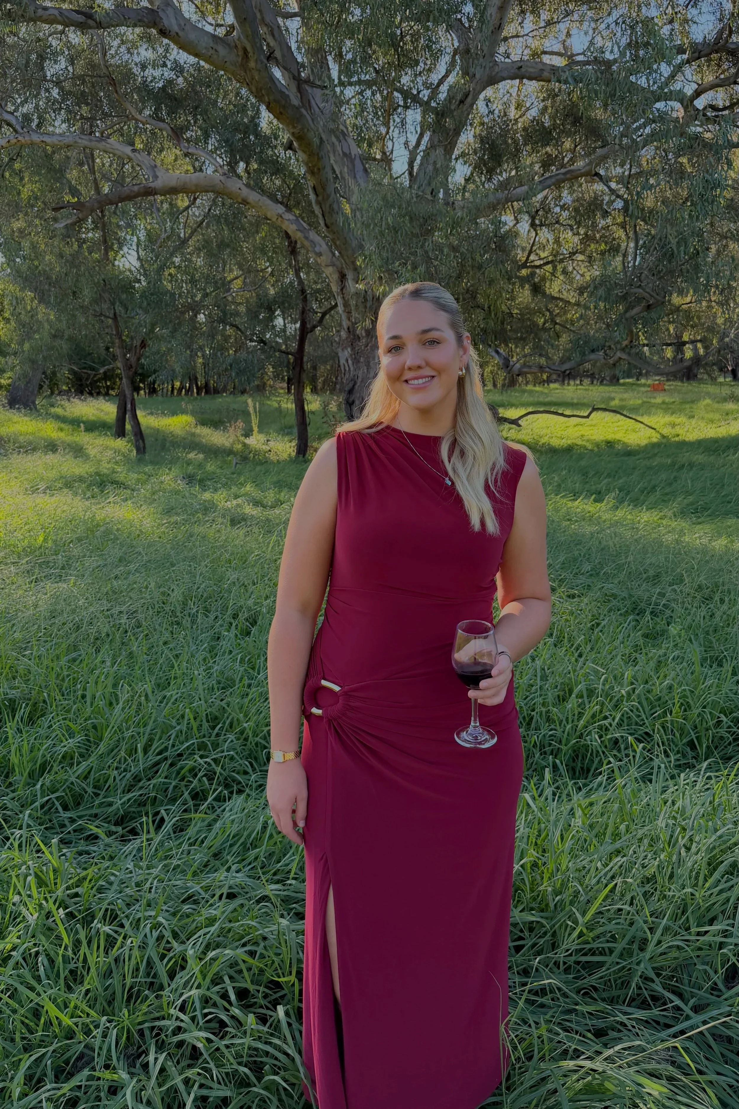 A woman in a sleeveless maroon evening gown stands in a grassy area near trees, holding a glass of red wine and smiling at the camera.