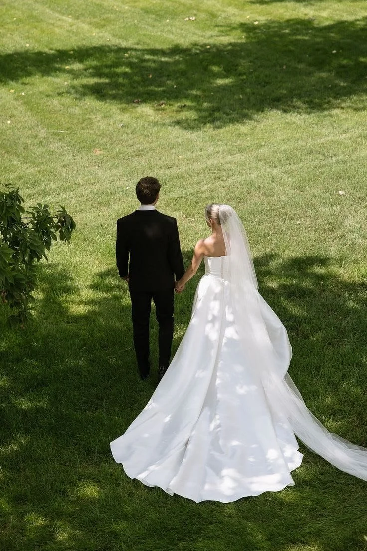A bride and groom walking hand in hand on a grassy outdoor area, seen from behind, during their wedding.