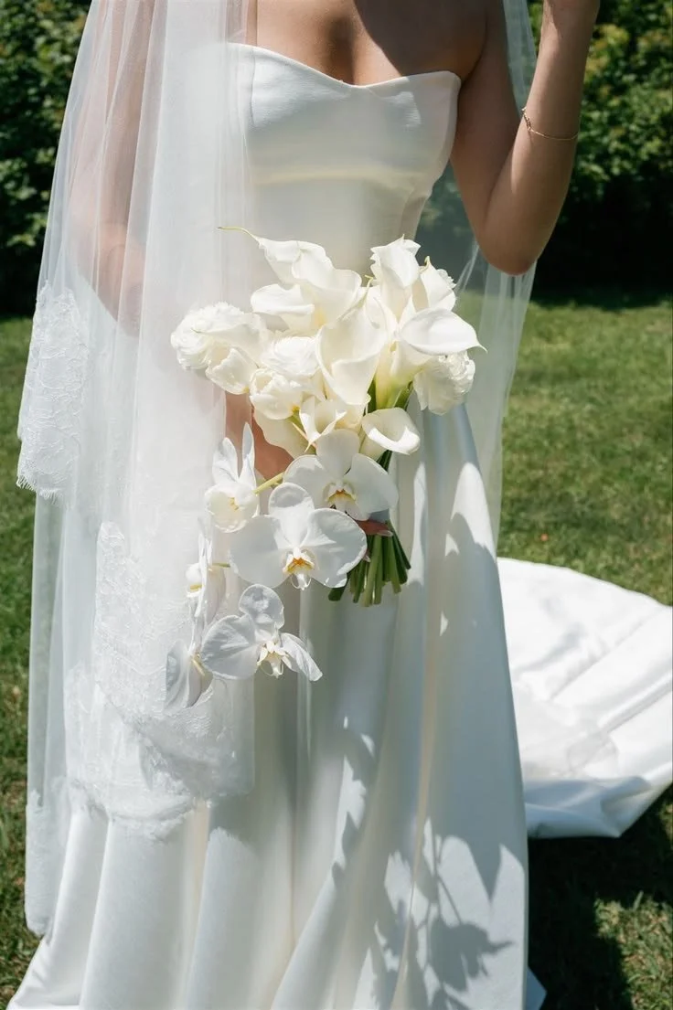 Close-up of a bride holding a bouquet of white orchids and calla lilies, wearing a white wedding dress with a sheer veil, standing on grass outdoors.