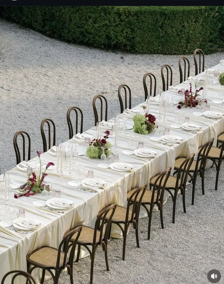 Long dining table set outdoors with white tablecloths, decorated with floral centerpieces, glassware, and napkins, surrounded by wooden chairs with woven seats.