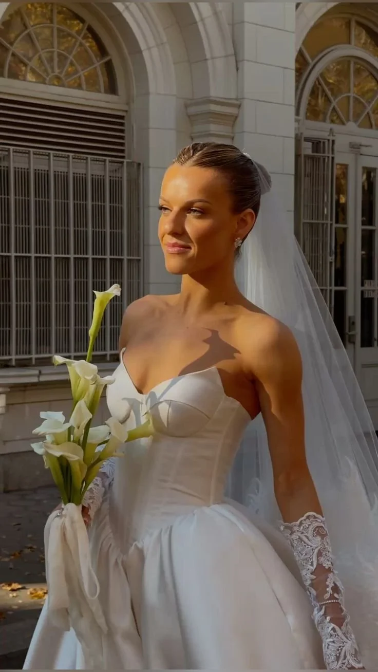 A bride in a white wedding gown holding a bouquet of calla lilies, standing outside a building with arched windows and classical architecture.