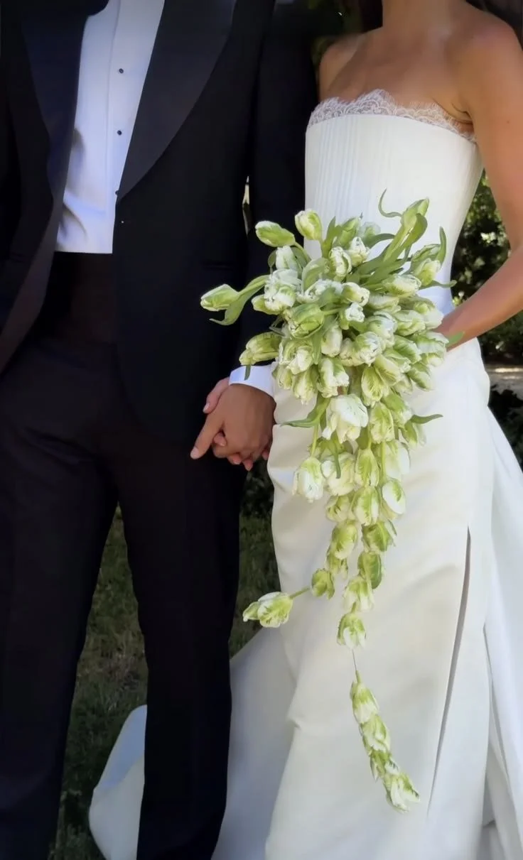 A bride and groom holding hands, with the bride in a white strapless wedding dress and holding a cascading bouquet of white and green flowers, standing outdoors.