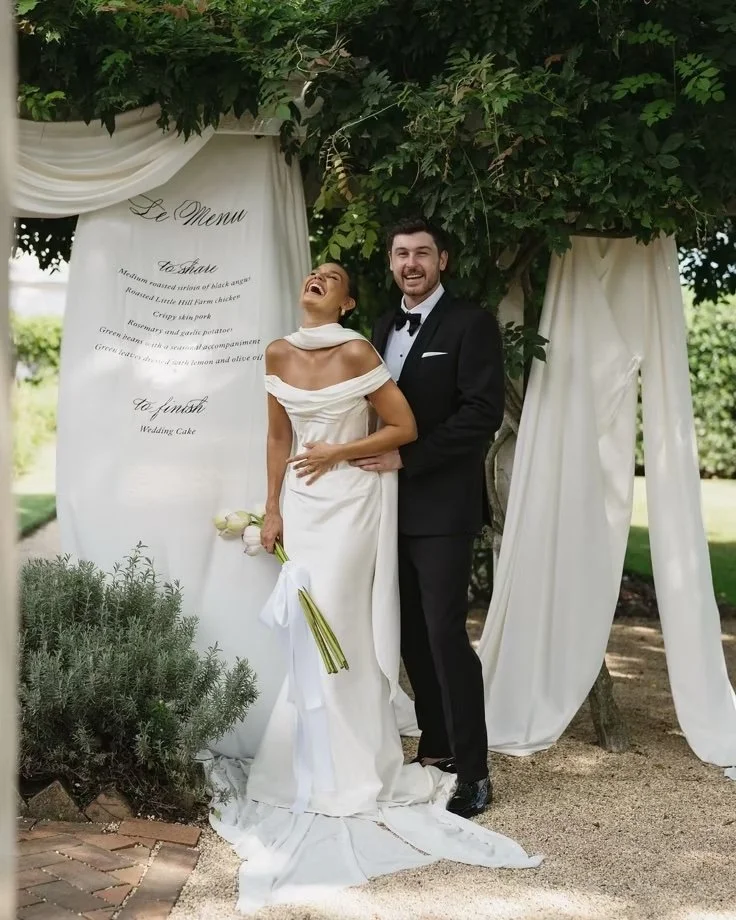 A bride and groom laughing together during their wedding ceremony outdoors, with a white draped backdrop and greenery.