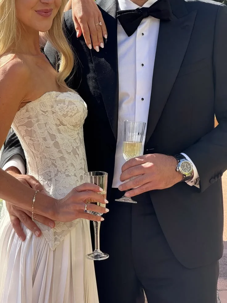 A bride and groom at a wedding celebration, holding glasses of champagne, with the bride wearing a lace dress and jewelry, and the groom in a tuxedo with a watch.