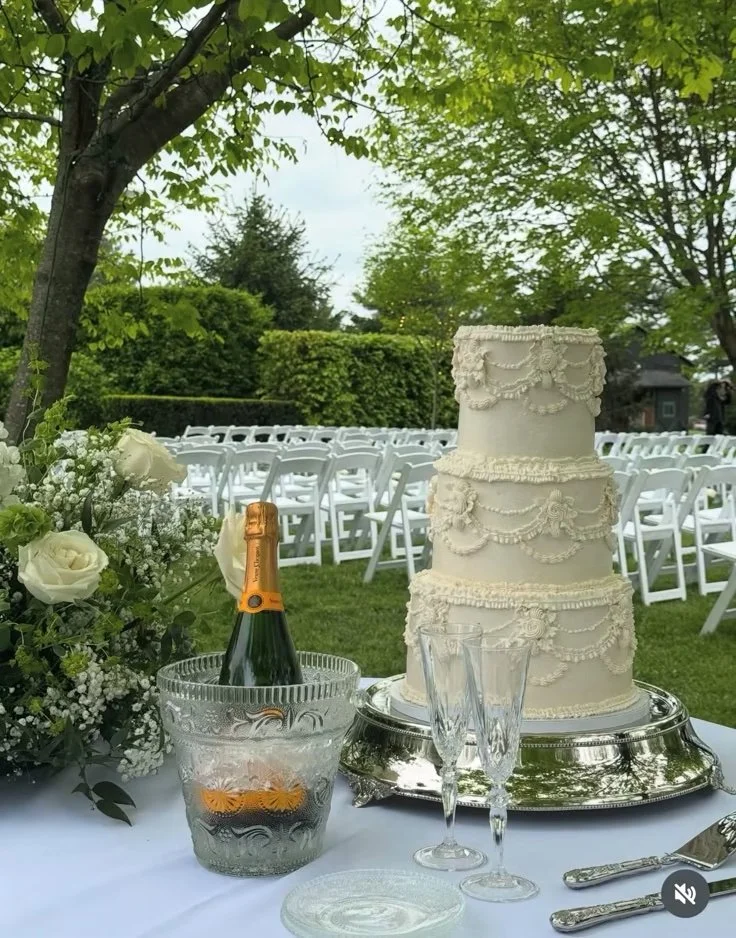 A four-tier wedding cake decorated with intricate icing designs sitting on a silver platter, placed on a table with champagne bottles in an ice bucket, champagne flutes, a silver cake knife, and a bouquet of white roses and baby's breath at an outdoo
