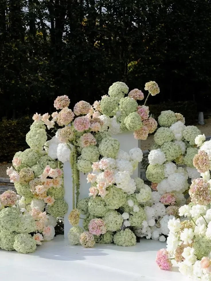 Decorative floral arrangement with white, pale pink, and light green hydrangeas and roses on a white table outdoors.