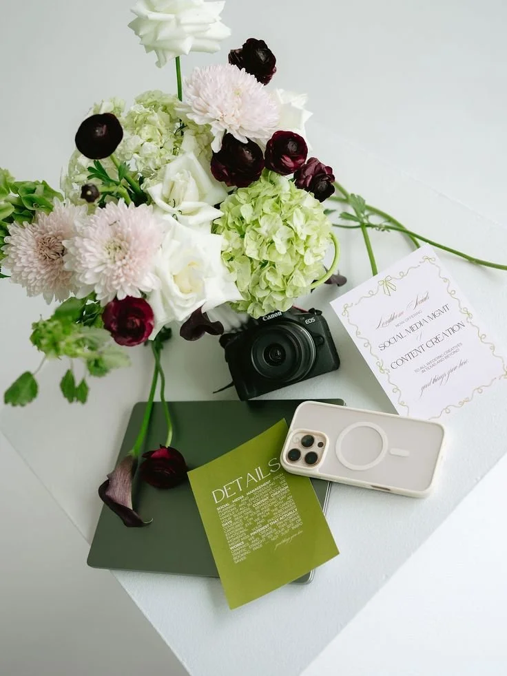 Flat lay of a bouquet of white, pink, and dark red flowers, a Canon camera, a smartphone with a pop socket, a green file or booklet with the text 'Details,' and a white card with gold decorative borders and the text 'Presentation for Social Media Mgm
