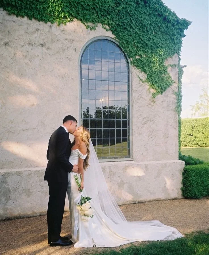 A newlywed couple sharing a kiss in front of a large window with green ivy on a stone building during sunset.