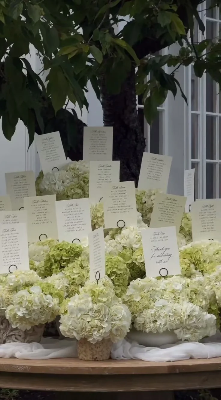 White hydrangea flower arrangements with cards on a table, a tree in the background.