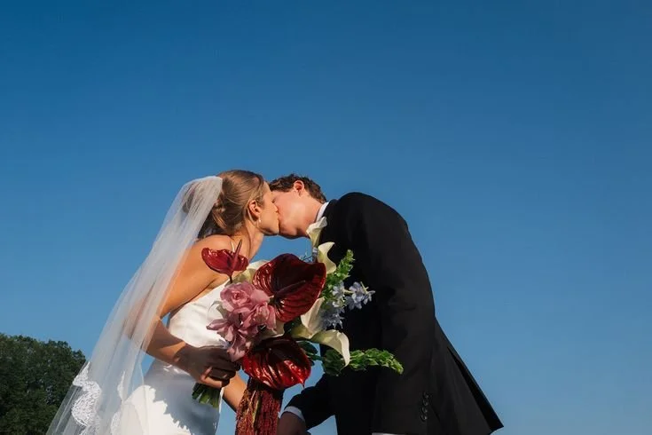 A bride and groom share a kiss outdoors, with the bride holding a large bouquet of flowers, against a clear blue sky.