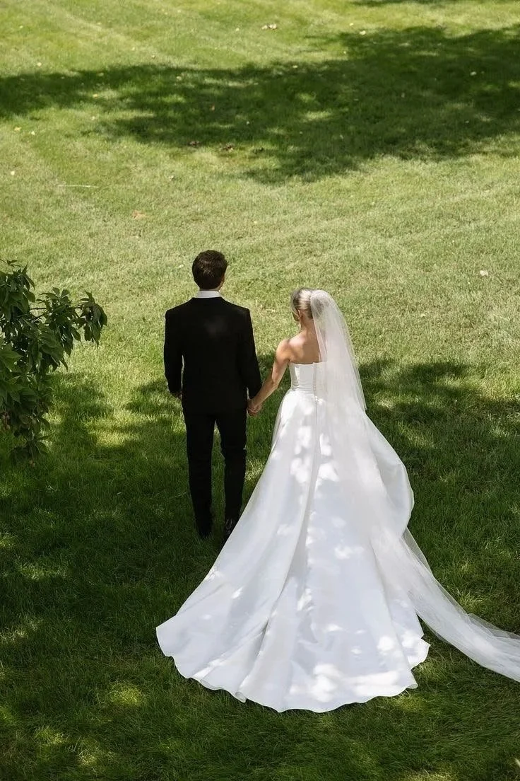 A bride and groom holding hands walk on grass during a wedding ceremony outdoors, with trees casting shadows in the background.