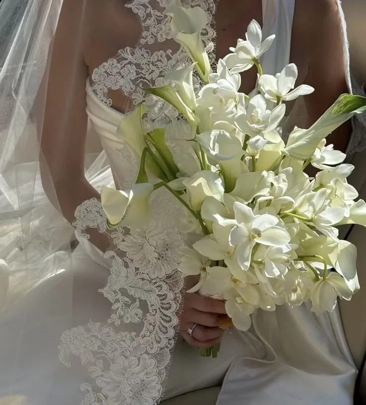 A bride in a white wedding dress with lace details, holding a bouquet of white flowers, including calla lilies and orchids.
