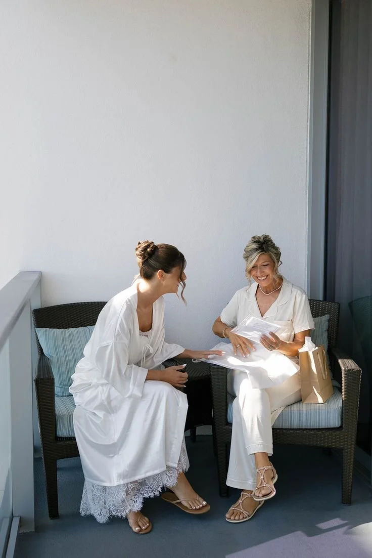 Two women sitting on a wicker sofa, smiling and looking at papers. One woman is wearing a white dress with lace details, and the other is wearing a white outfit with sandals. They are on a balcony with a plain wall behind them.