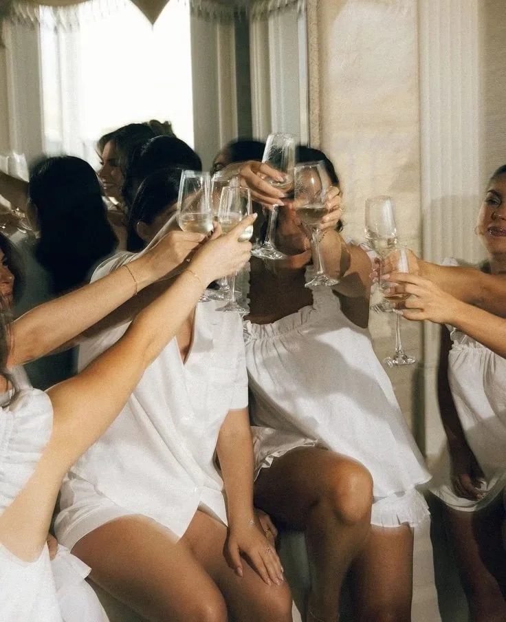 Several women wearing white robes are sitting together, raising their glasses of white wine in a toast during a celebration or gathering.