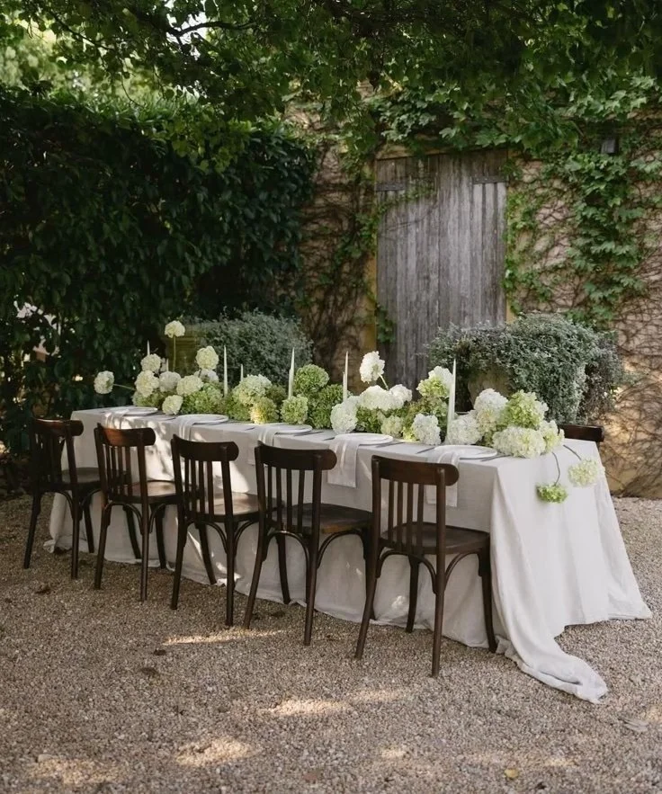 A long outdoor dining table decorated with white tablecloth, white floral arrangements, and tall white candles, set in a garden area with gravel ground, surrounded by lush green foliage and a weathered wooden wall.