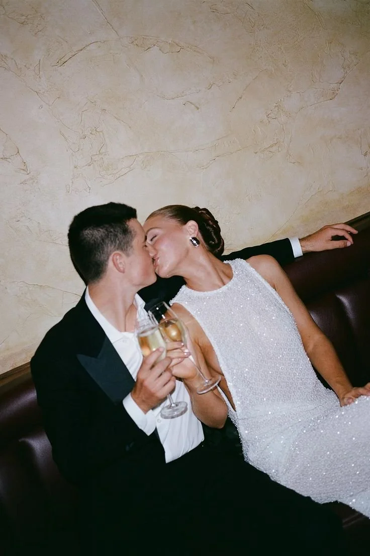 A couple dressed in formal attire sharing a kiss while holding glasses of champagne, sitting on a dark leather bench against a beige wall.