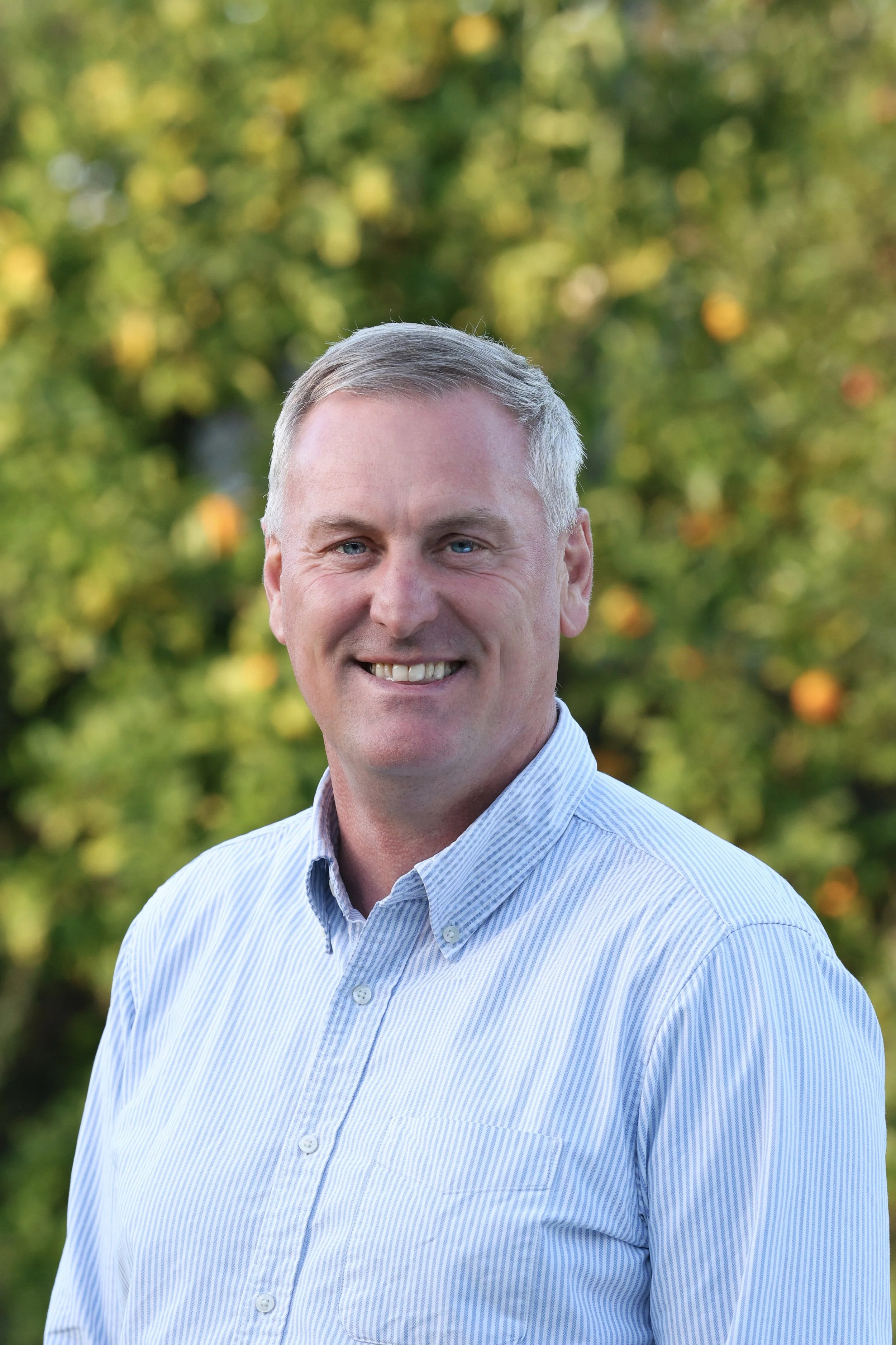 A smiling man with short gray hair wearing a light blue, white-striped button-up shirt, standing outdoors with blurred green foliage and orange fruits in the background.