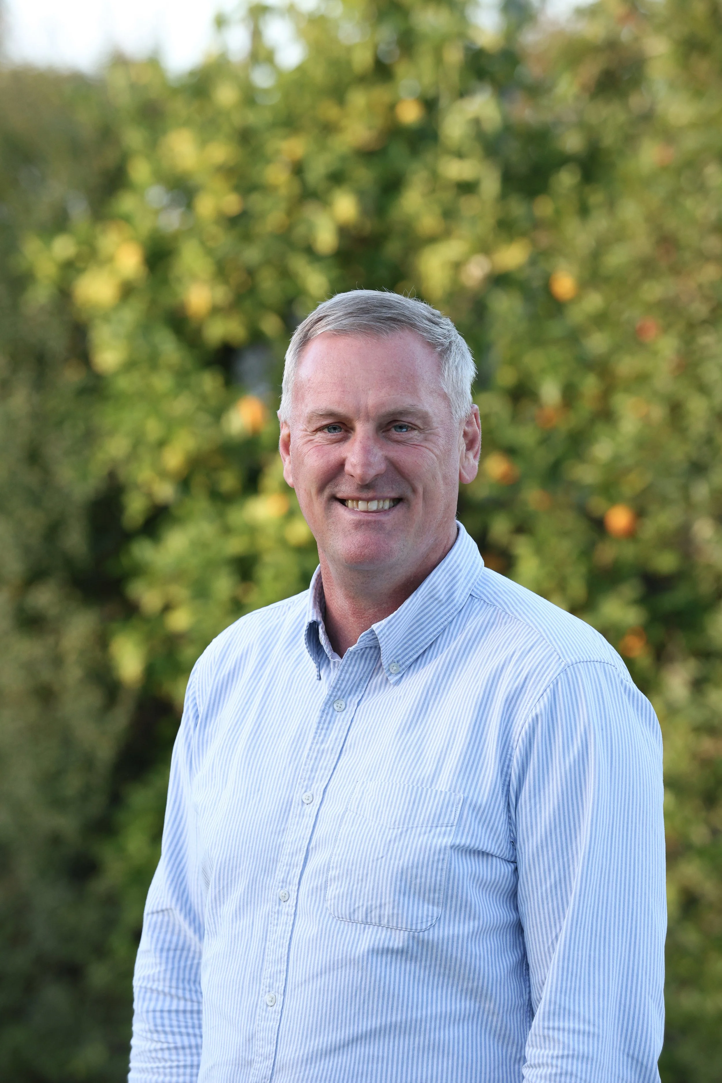 Mark Scanlon Next Transition Group A middle-aged man with short gray hair, wearing a light blue, striped button-up shirt, smiling outdoors with blurred green trees and orange fruits in the background.