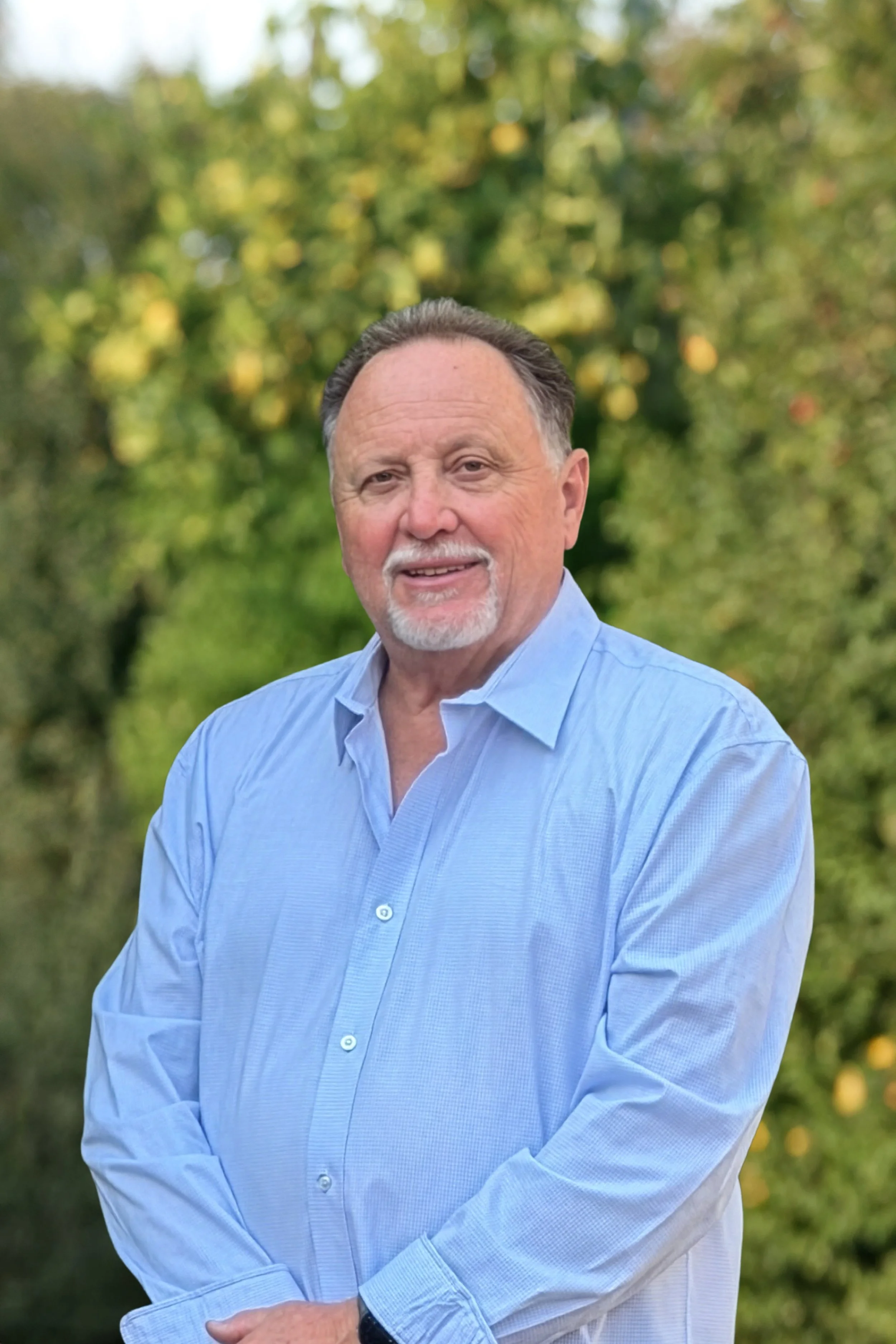 A smiling older man with a beard and mustache, wearing a light blue button-down shirt, standing outdoors with blurred green trees in the background.