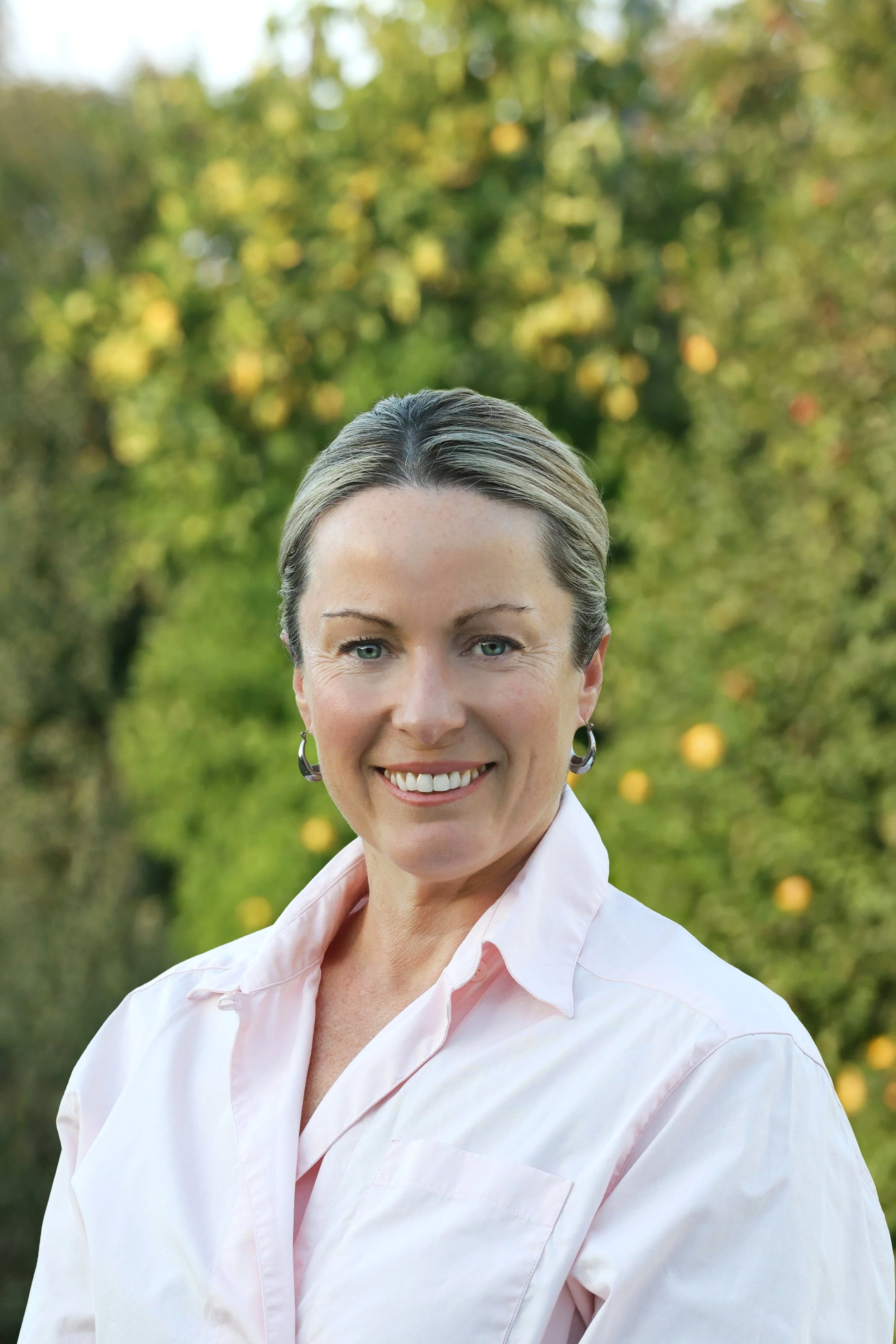 A woman with short gray hair and blue eyes smiling outdoors, wearing a light pink shirt, with a blurred background of green and yellow foliage.