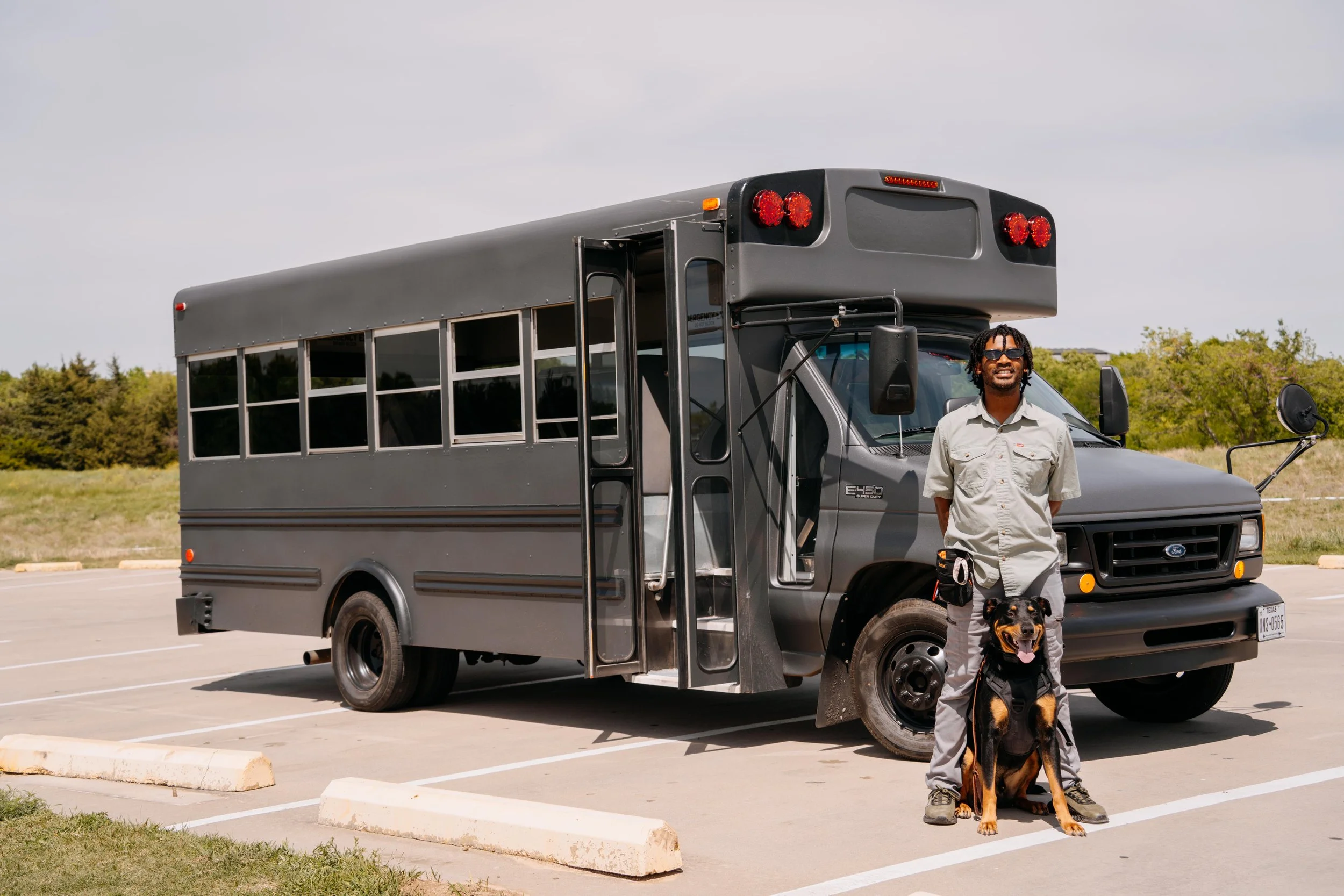 Man standing in a parking lot beside a dog and a black bus, with trees and a cloudy sky in the background.