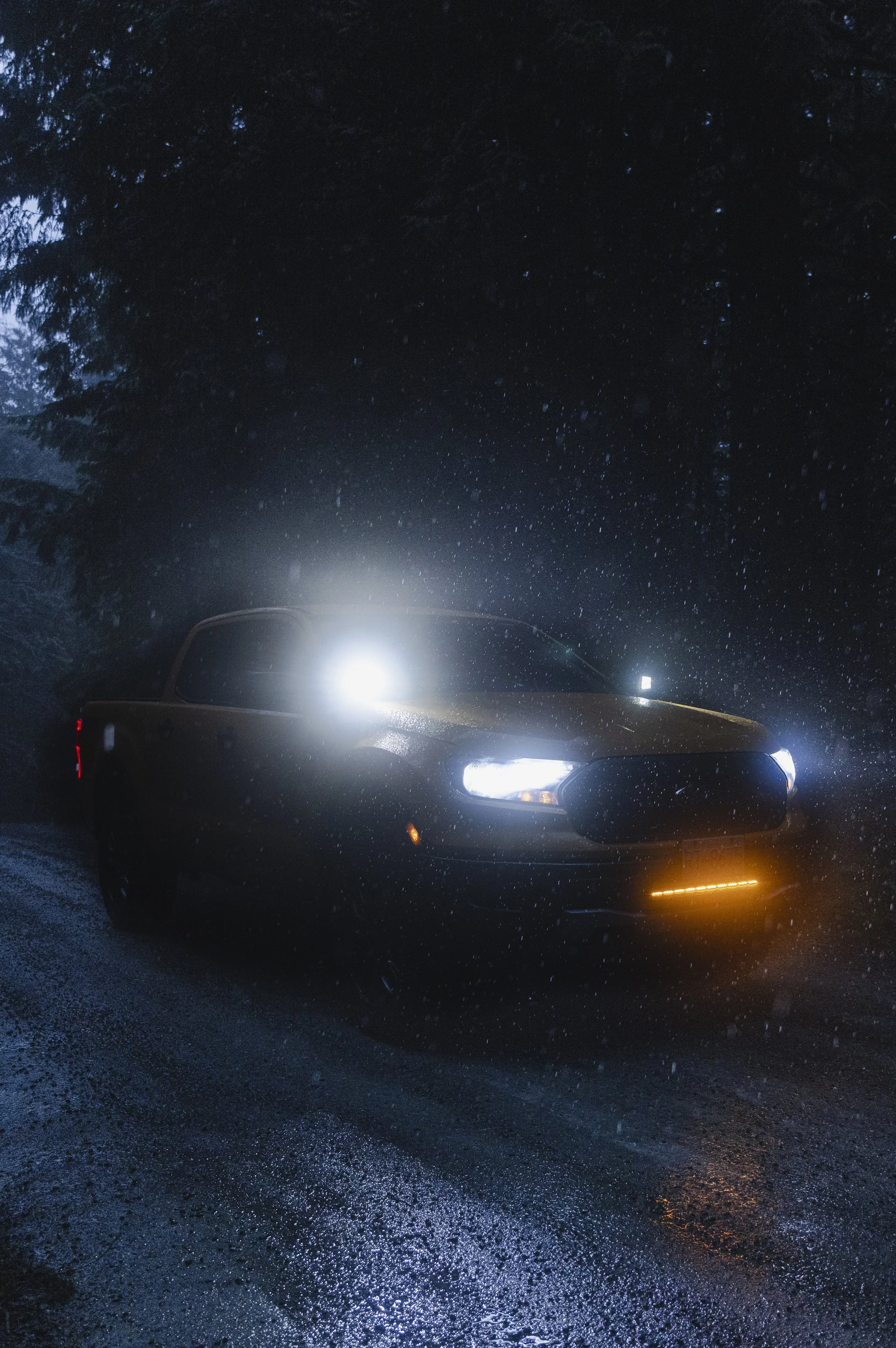 A black car with headlights on, parked on a wet road at night, with trees in the background and rain falling.