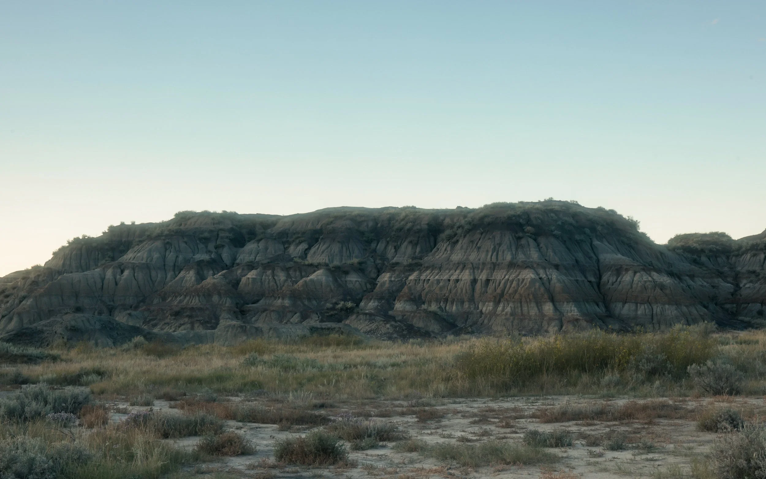Photo of a desert landscape with layered rock formations and sparse vegetation under a clear sky.