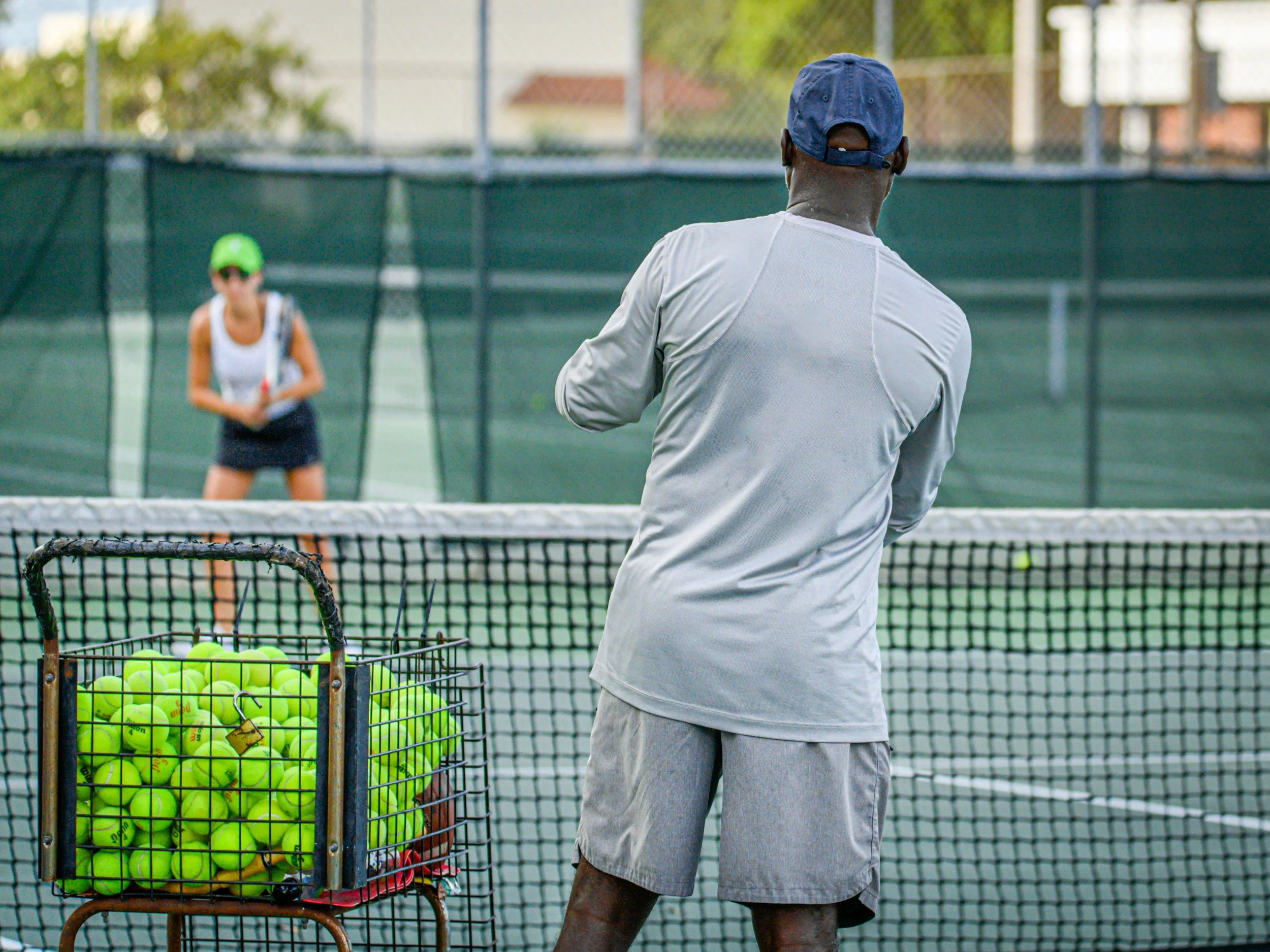 A man in a gray shirt and shorts stands on a tennis court, facing a woman who is preparing to hit a tennis ball. There is a basket filled with tennis balls in the foreground.