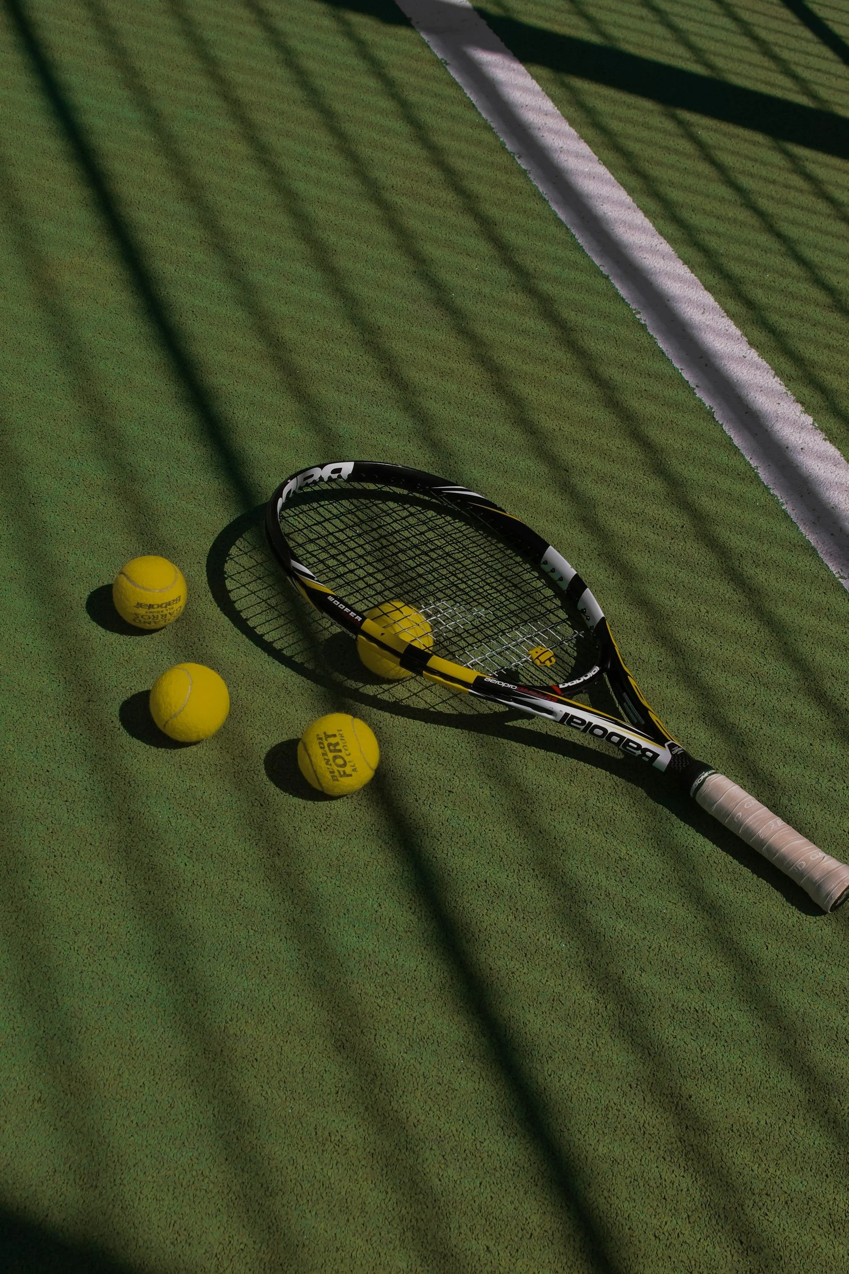 A tennis racket and four yellow tennis balls on a green outdoor tennis court with a white boundary line, cast in warm sunlight with shadows.